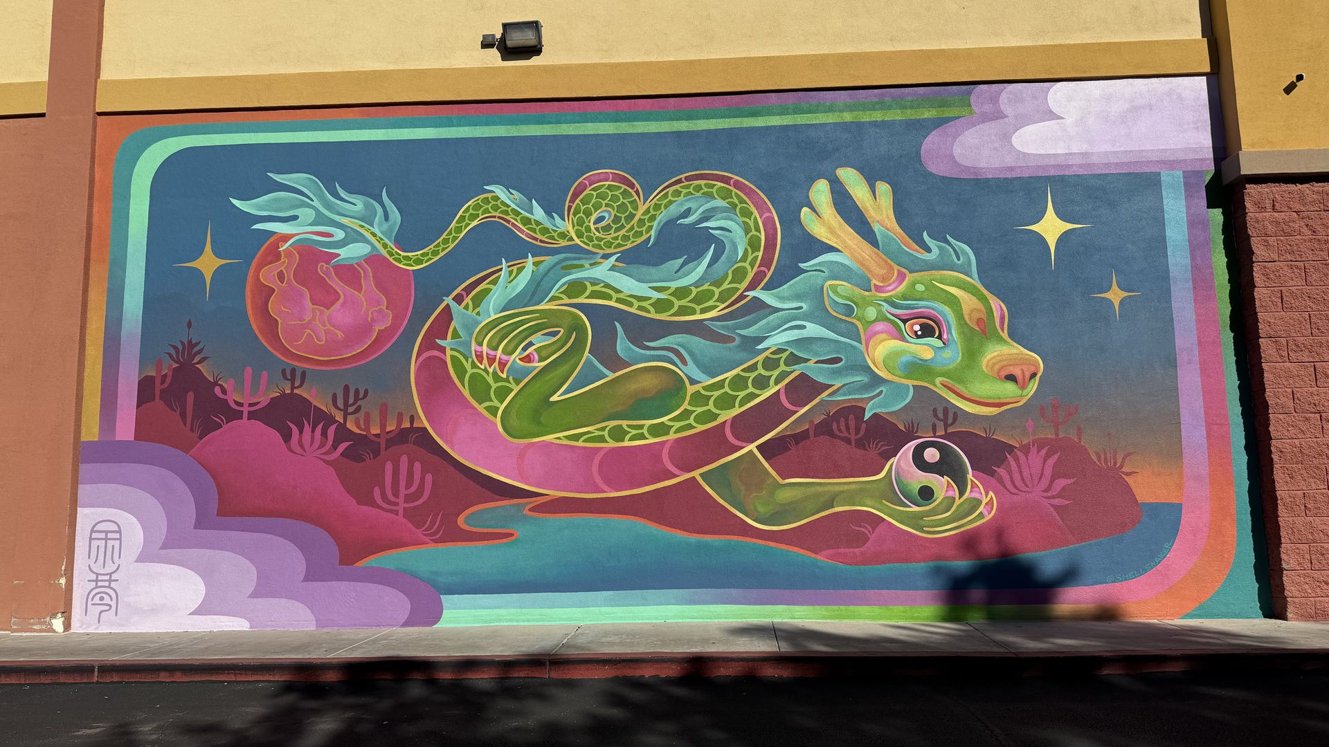 Colorful mural of a green, serpentine dragon with blue flames, holding a black and white yin-yang orb, flying over a pink desert landscape with cacti, stars, and a red sun in the background.