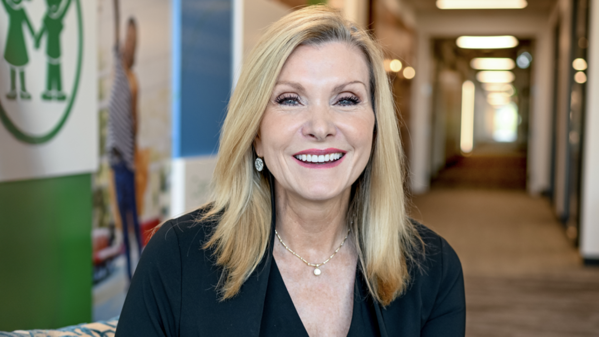 Smiling blonde woman in a black blazer sits in a bright office hallway, wearing a delicate necklace and pearl earrings. A green poster is visible on the left.