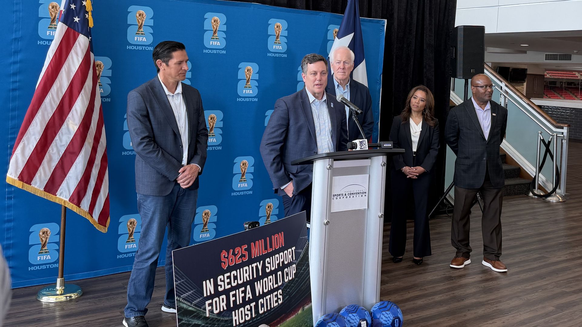 Press conference with suited officials at a podium in front of a blue FIFA Houston backdrop; a U.S. flag on the left and a banner about $625 million in security for FIFA World Cup host cities.