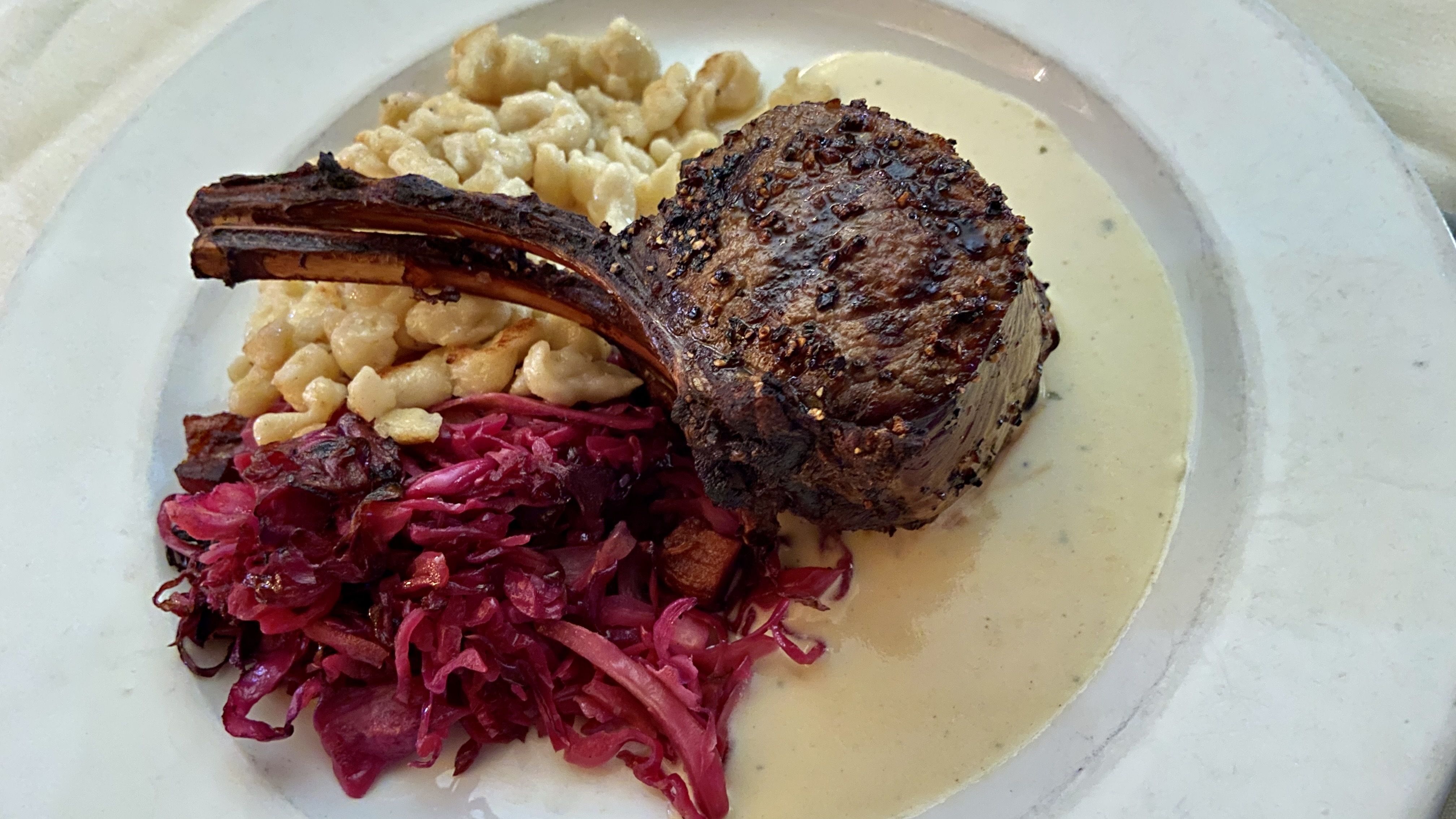 A plate featuring a seasoned and grilled lamb chop with a rib bone, placed on a creamy white sauce. Accompanied by a serving of spaetzle pasta and a side of red cabbage. The dish is arranged on a white plate.