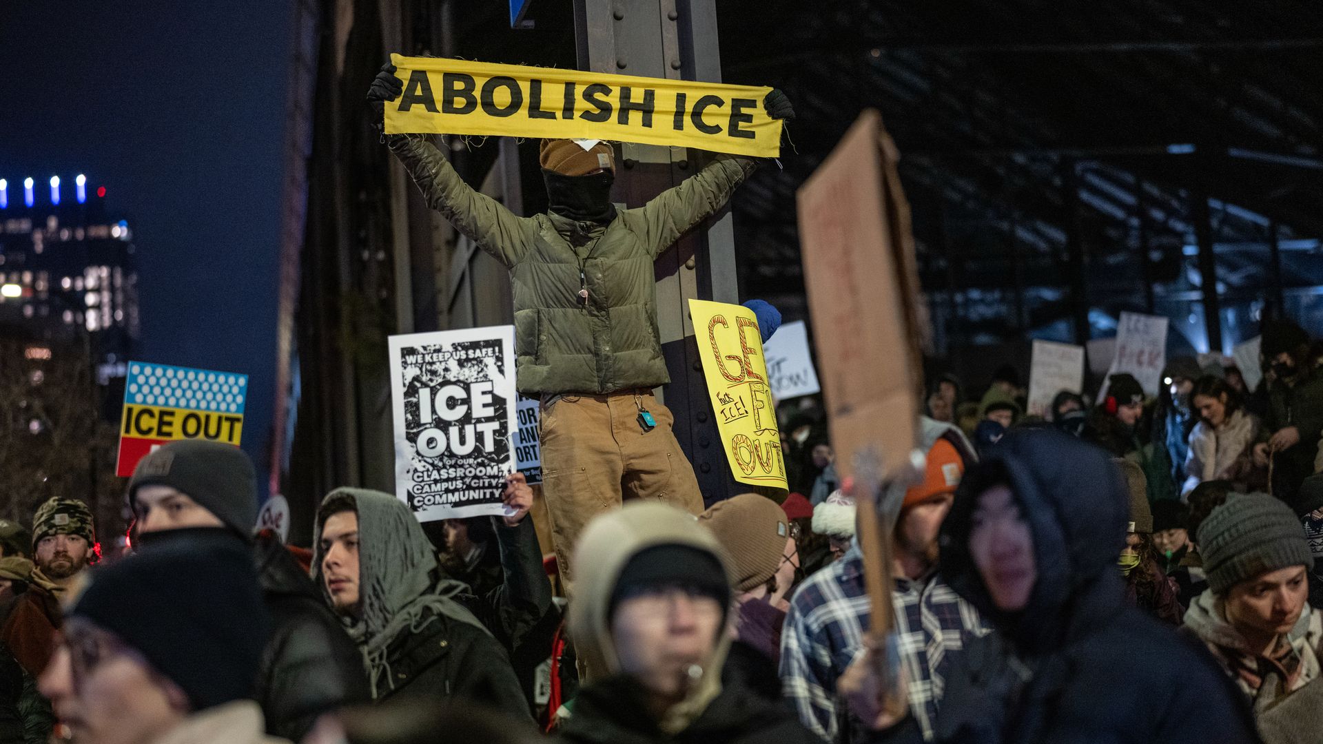 Protesters bundled in winter clothes hold signs at night, including a yellow banner stating "ABOLISH ICE" and others saying "ICE OUT" and "GET ICE OUT."