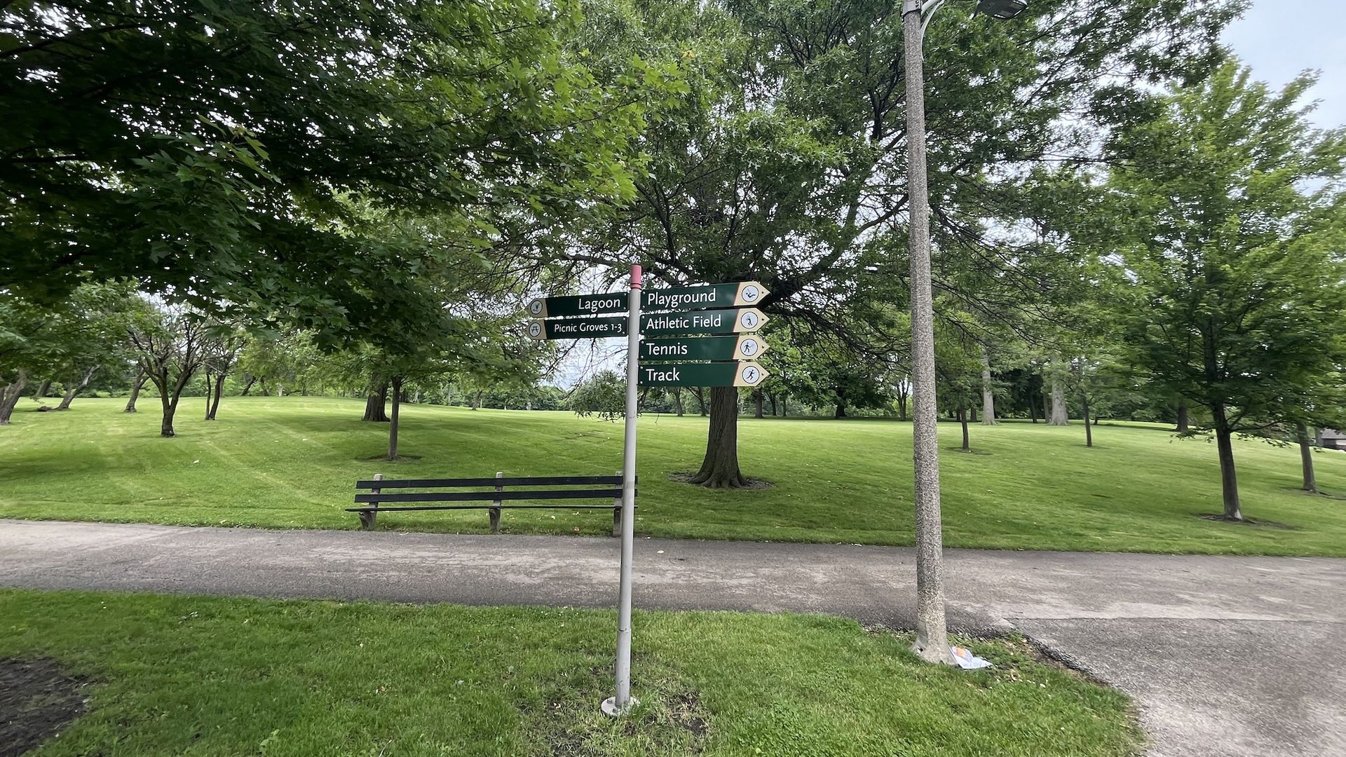 A park scene with a multi-directional sign near a bench and a tall street lamp. Green trees and grass surround the sign pointing to Lagoon, Playground, Picnic Groves, Athletic Field, Tennis, and Track.