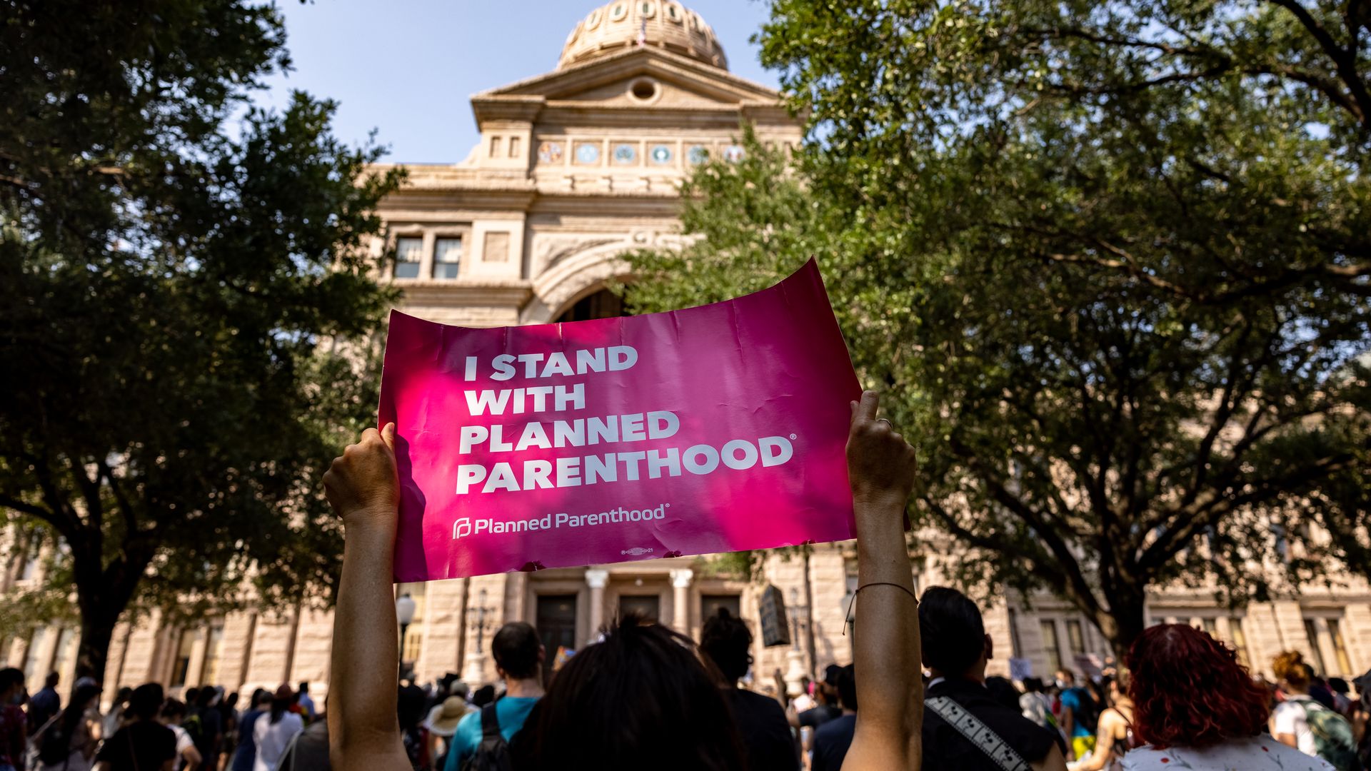 Photo of two arms holding up a pink sign that says "I stand with Planned Parenthood" in a crowd of pro-abortion protesters in front of the Texas state capitol