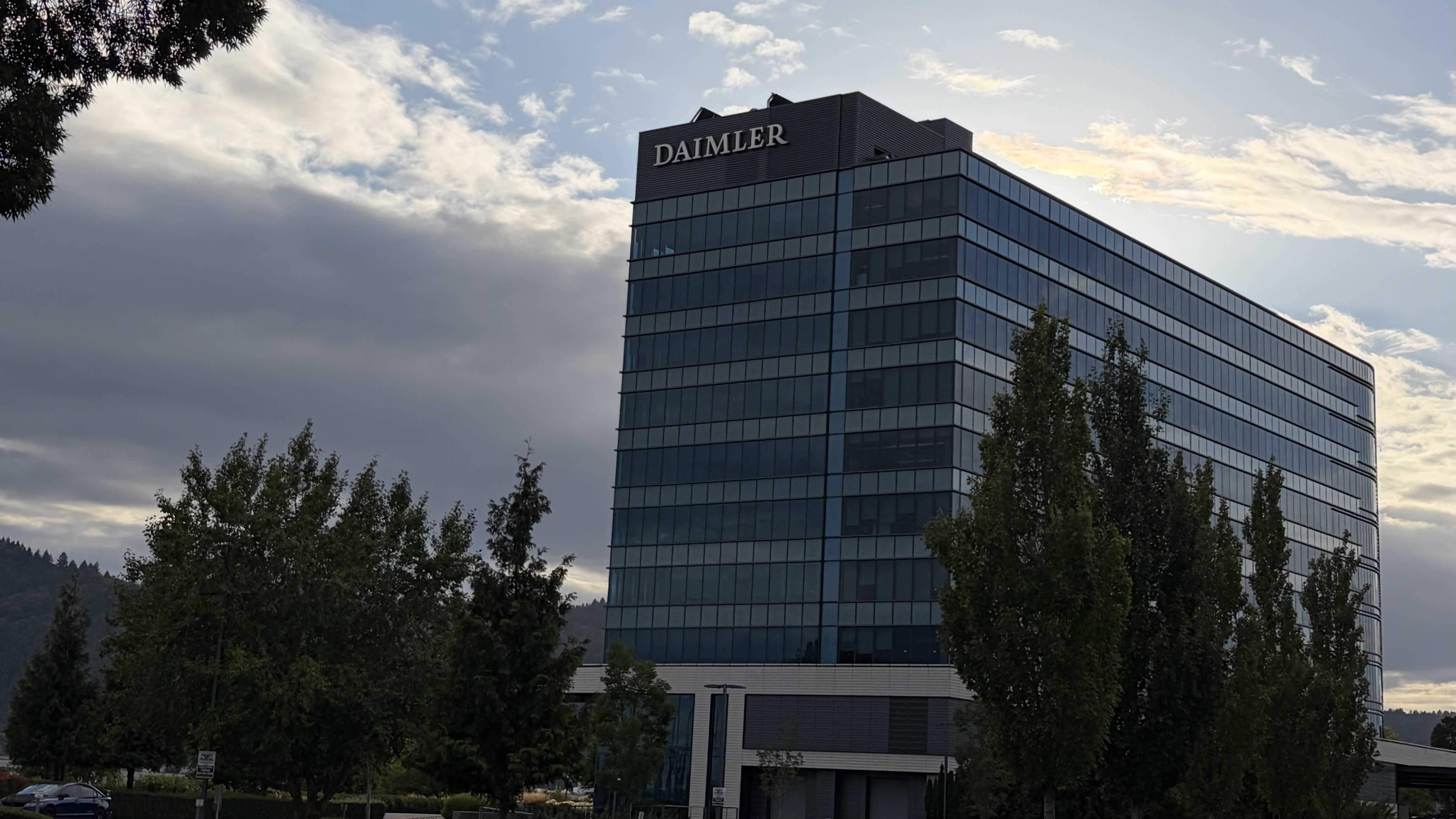 Modern glass office building with a DAIMLER sign atop, reflecting the blue sky. Foreground trees, a parking lot, and distant hills under a partly cloudy sky.