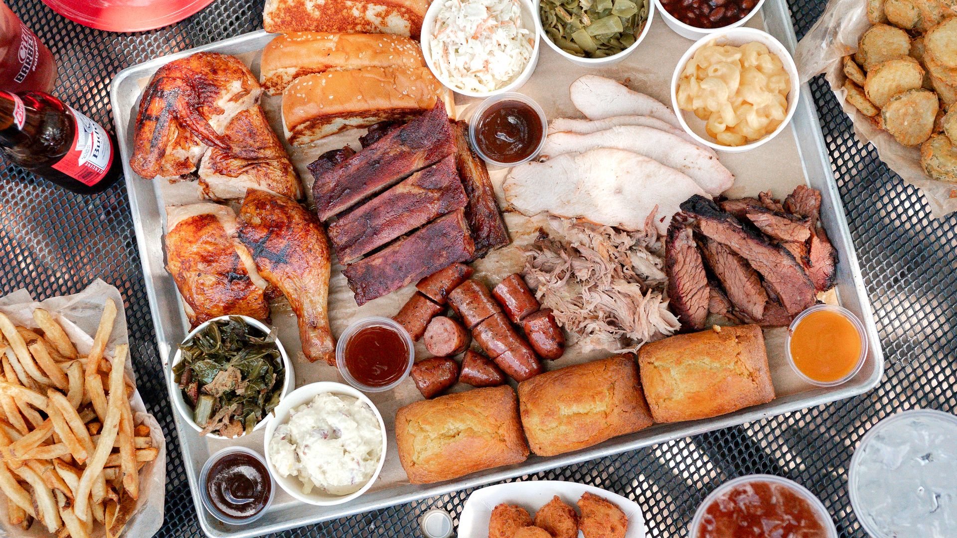 Tray of barbecue food including grilled chicken, ribs, sausage, turkey, pulled pork, brisket, cornbread, coleslaw, green beans, baked beans, mac and cheese, collard greens, fries, hush puppies, pickles, sauces, beer, and iced tea on a black mesh table.