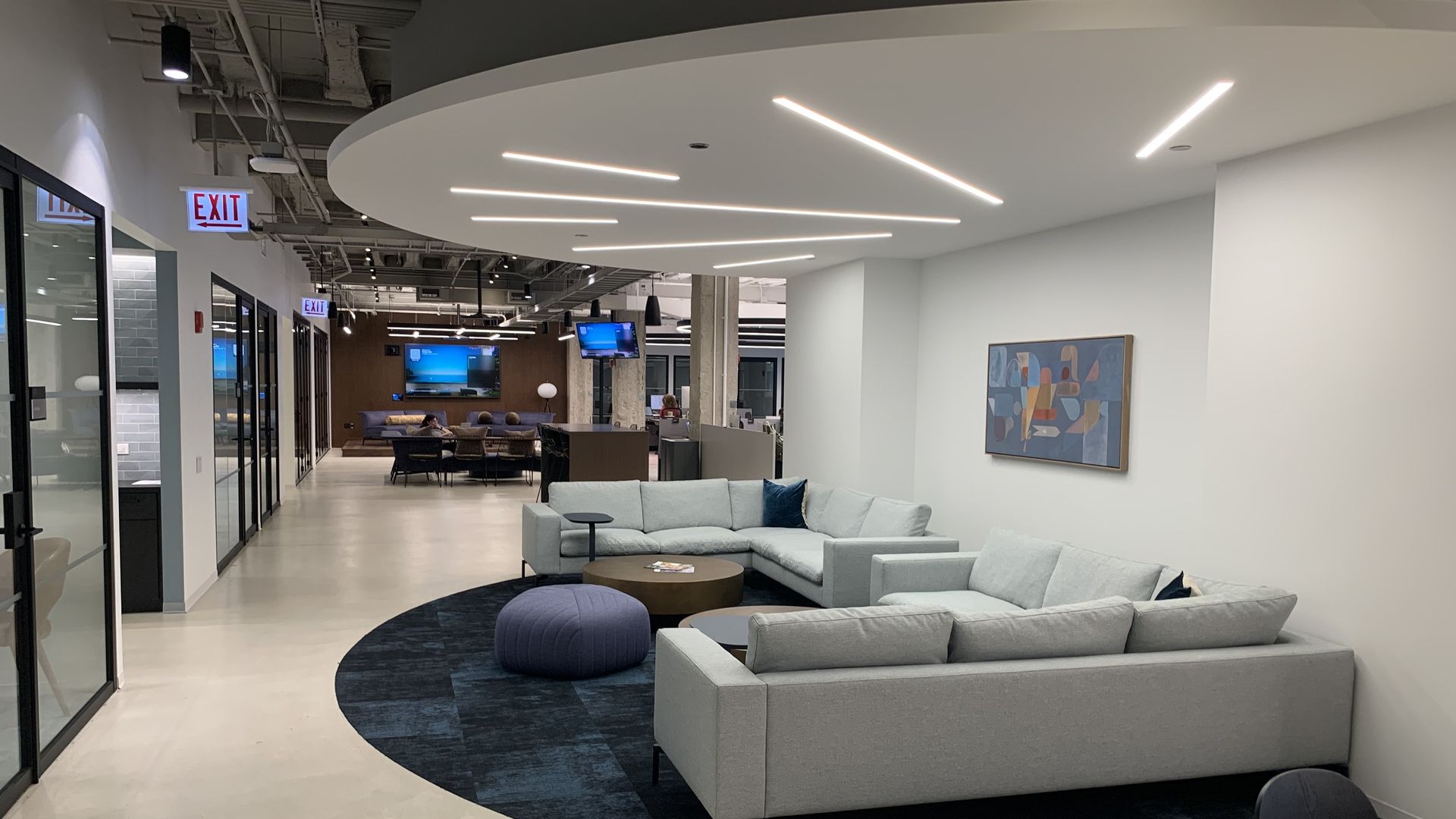 Modern office lounge with gray sectional sofas, blue pouf, dark round tables on a navy rug, abstract art on white walls, and bright linear ceiling lights.