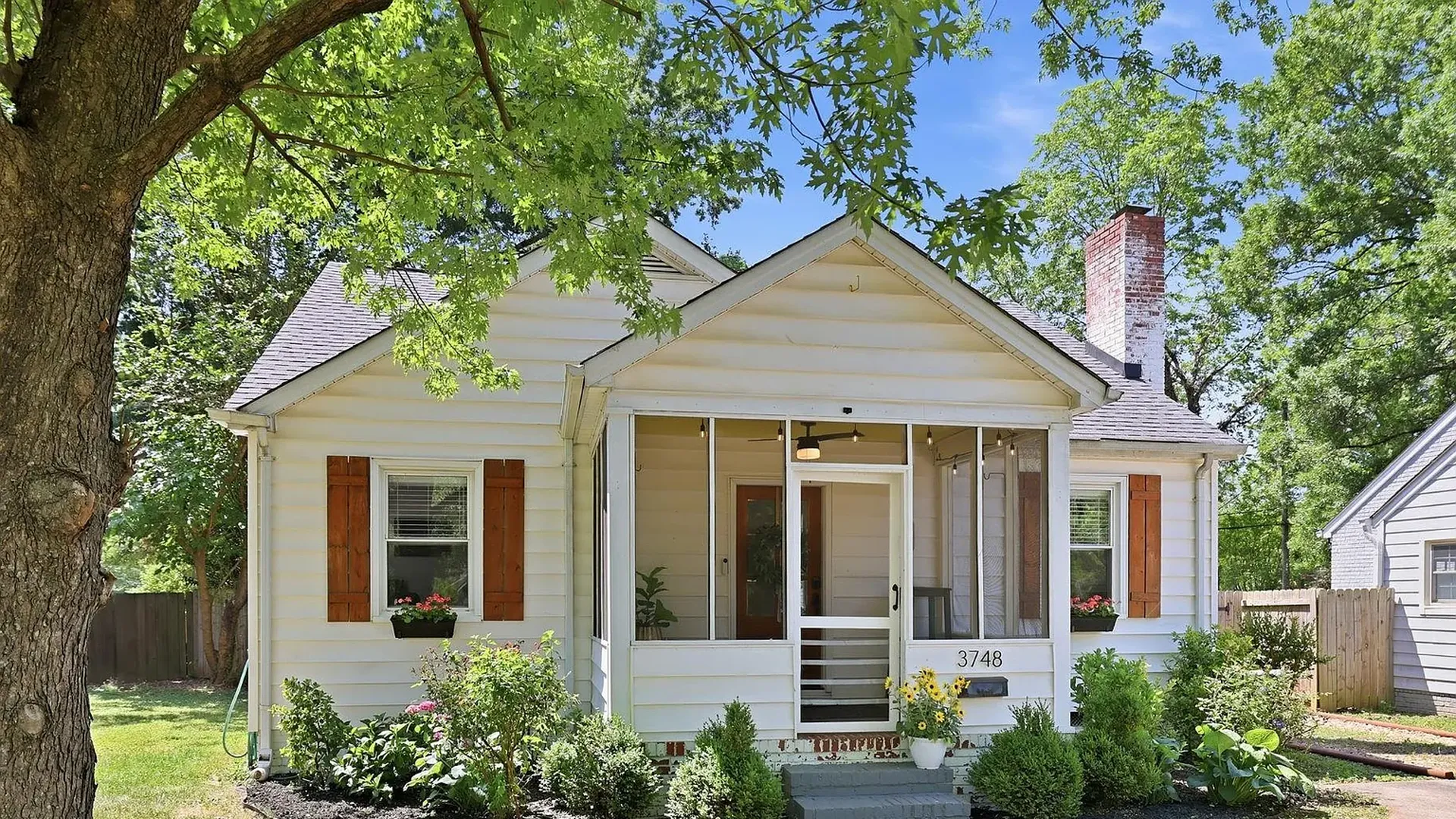 Small white house with a screened front porch, brown shutters, and a brick chimney. Front garden with flowers, a large tree in the foreground, and the number 3748 near the entry.