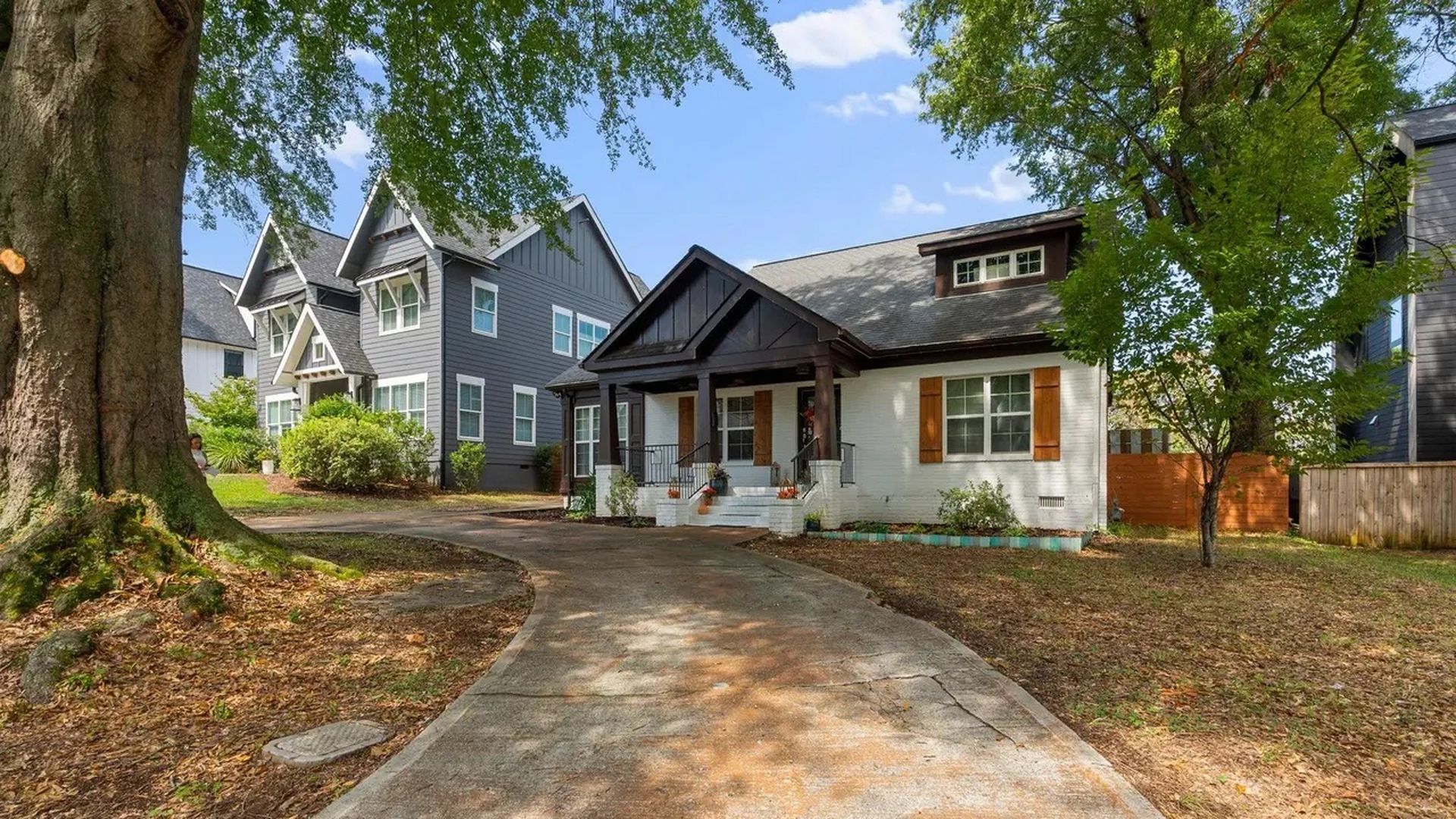 Curved concrete driveway leading to a white brick house with dark wooden porch and brown shutters, surrounded by large trees and neighboring grey houses under a blue sky.