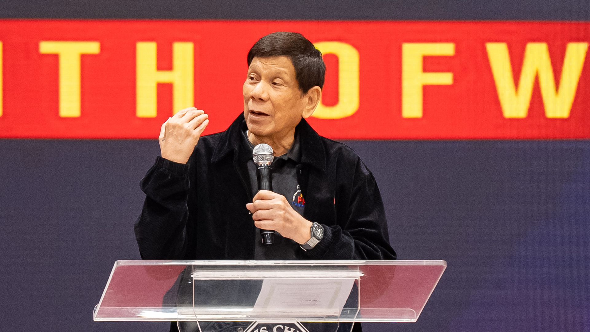 Former Philippine President Rodrigo Duterte, wearing a black jacket and shirt, gestures with his right hand while holding a mic in his left at a clear podium with the words "Jesus Christ" written on it.
