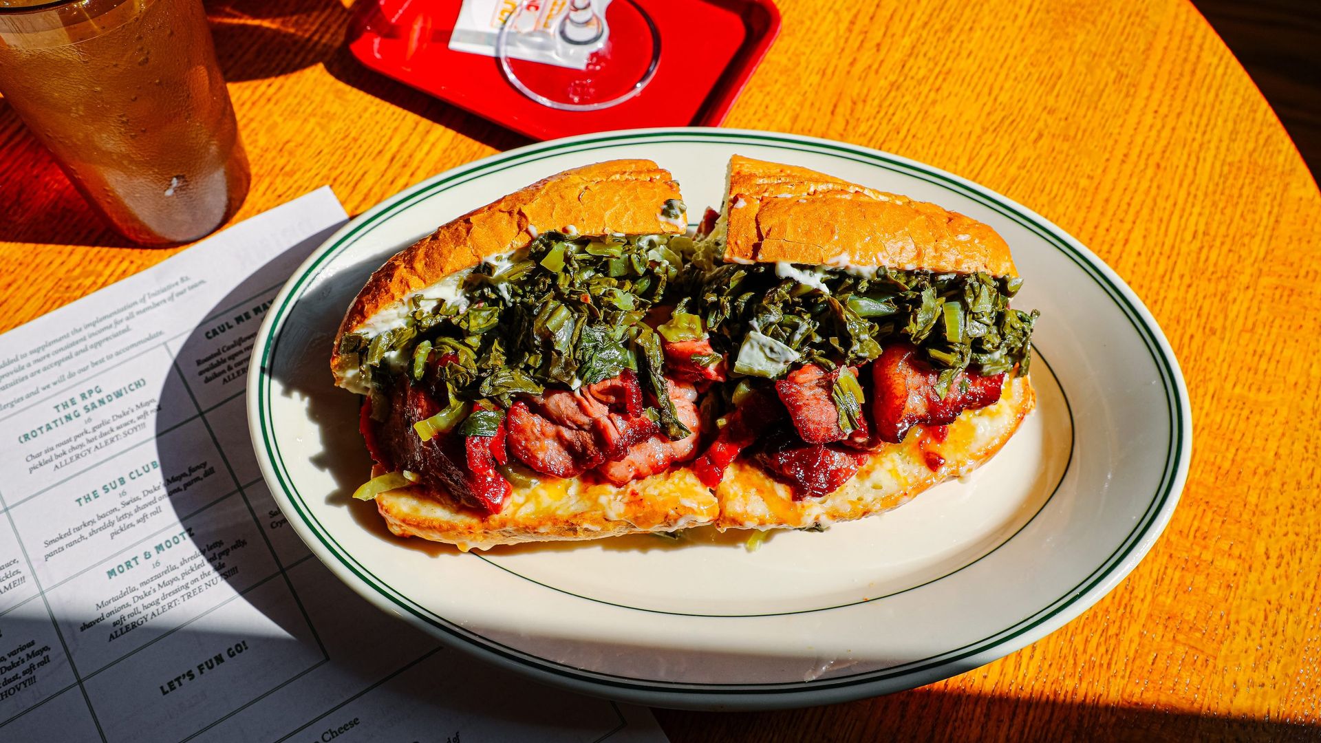 A sandwich filled with cooked greens, thick slices of grilled meat, and cheese on a white plate with a green rim on a wooden table. Nearby are a glass of iced tea and a red tray with a wine glass.