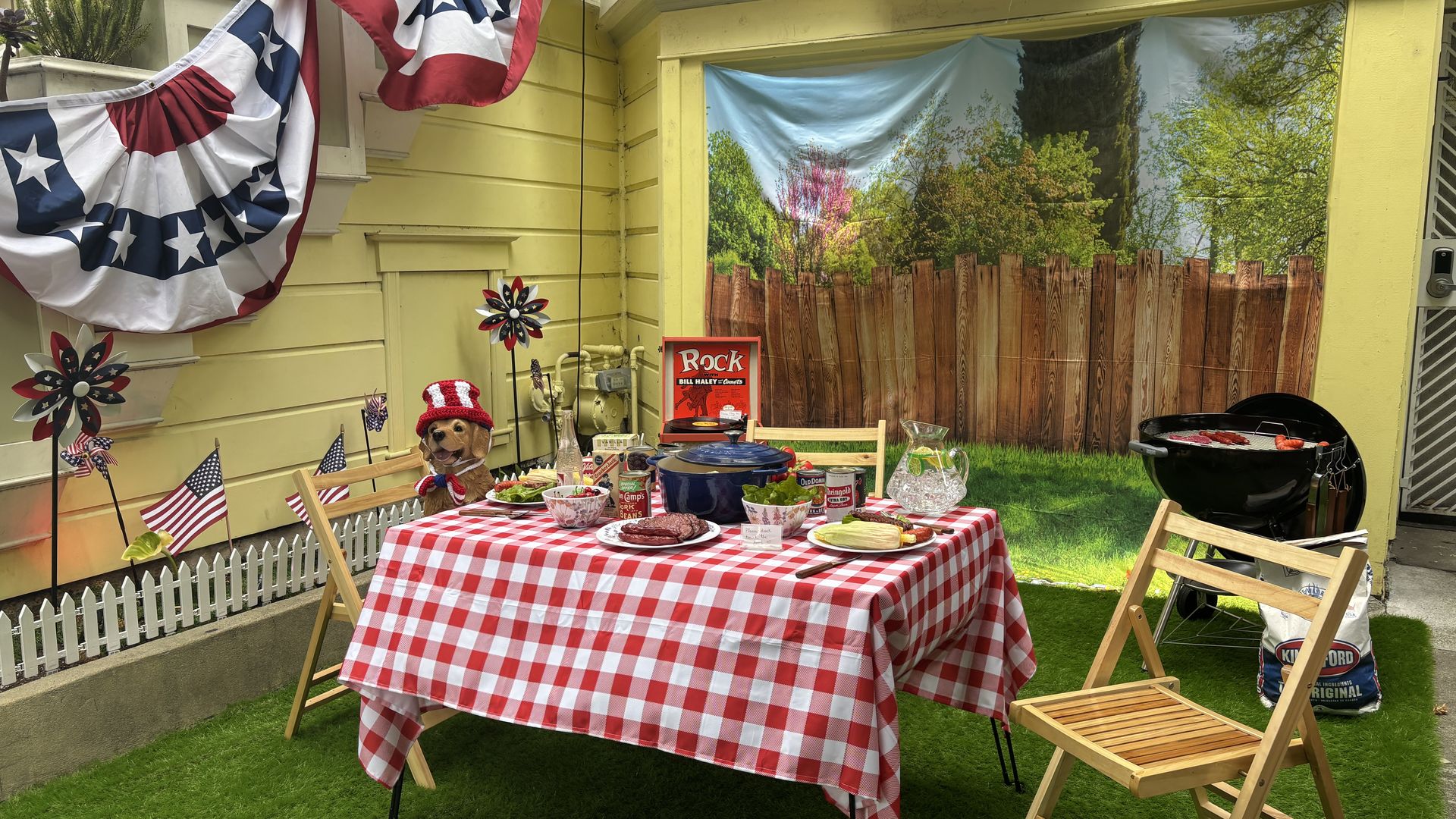 Patriotic backyard barbecue with a yellow house, red white blue bunting and flags, a red and white checkered tablecloth, grill on the right, and a table of food and drinks.