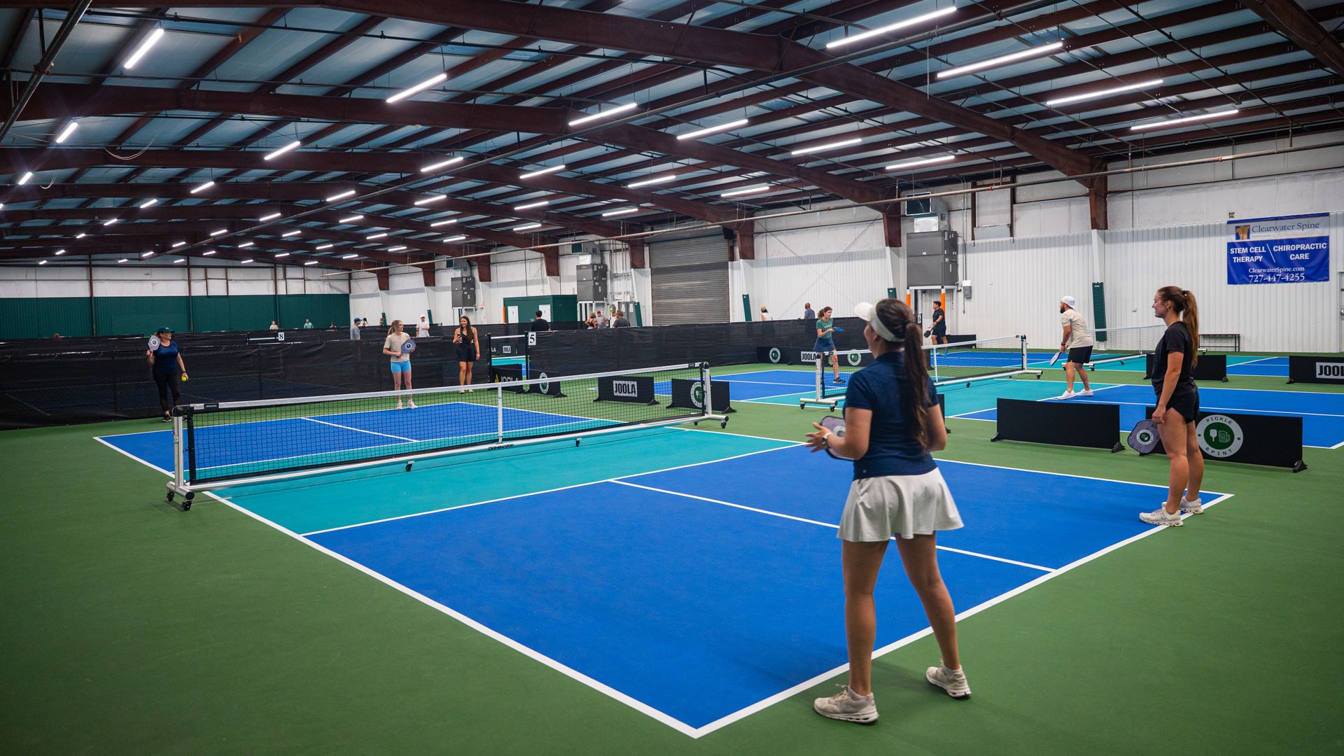 Indoor pickleball court with blue and green surfaces, several players holding paddles and balls, bright overhead lights, and a visible Chiropractic Care sign on the wall.