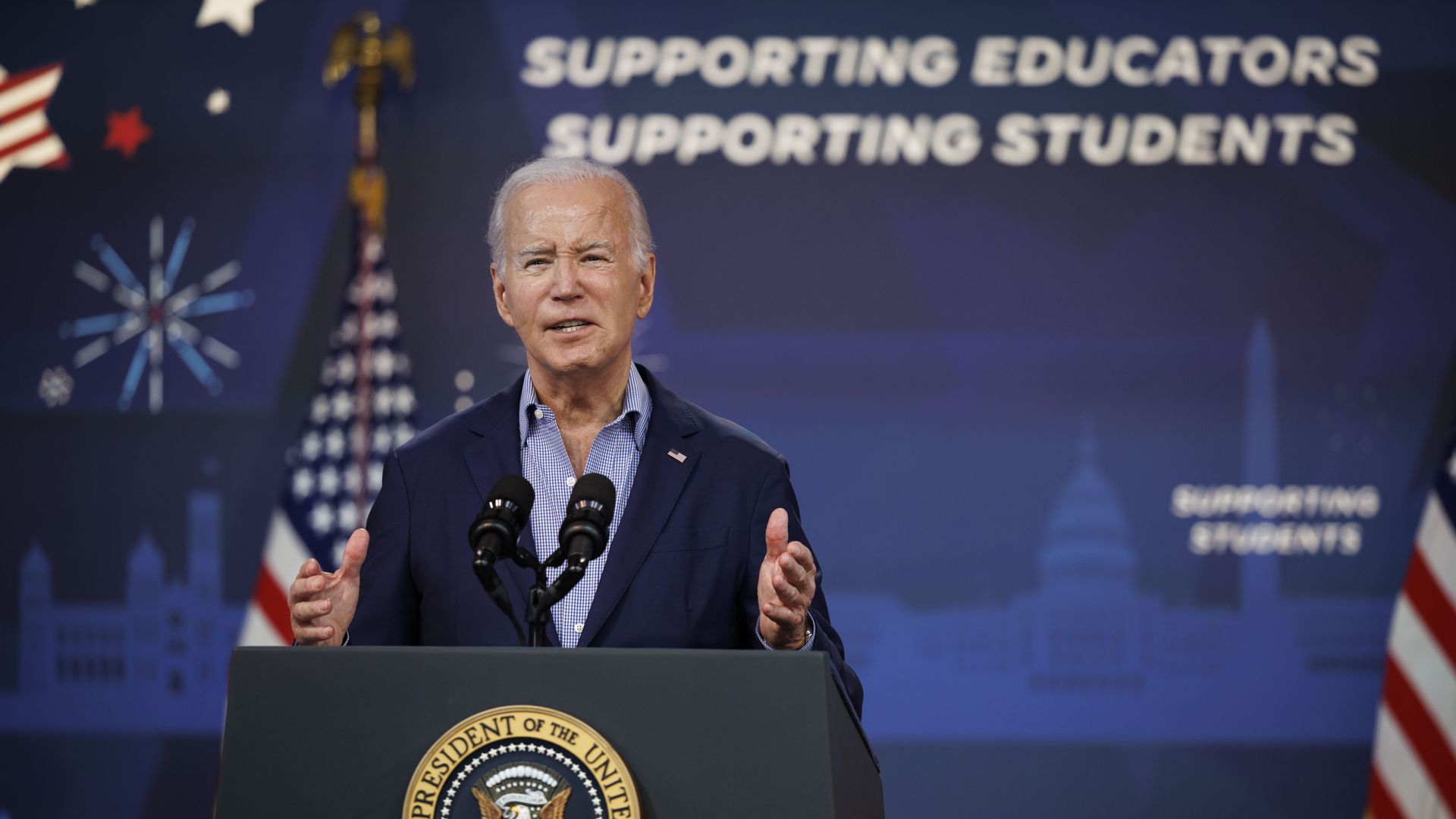 Joe Biden speaks at a lectern with hands raised. In the background, a sign reads "Supporting educators. Supporting students." 