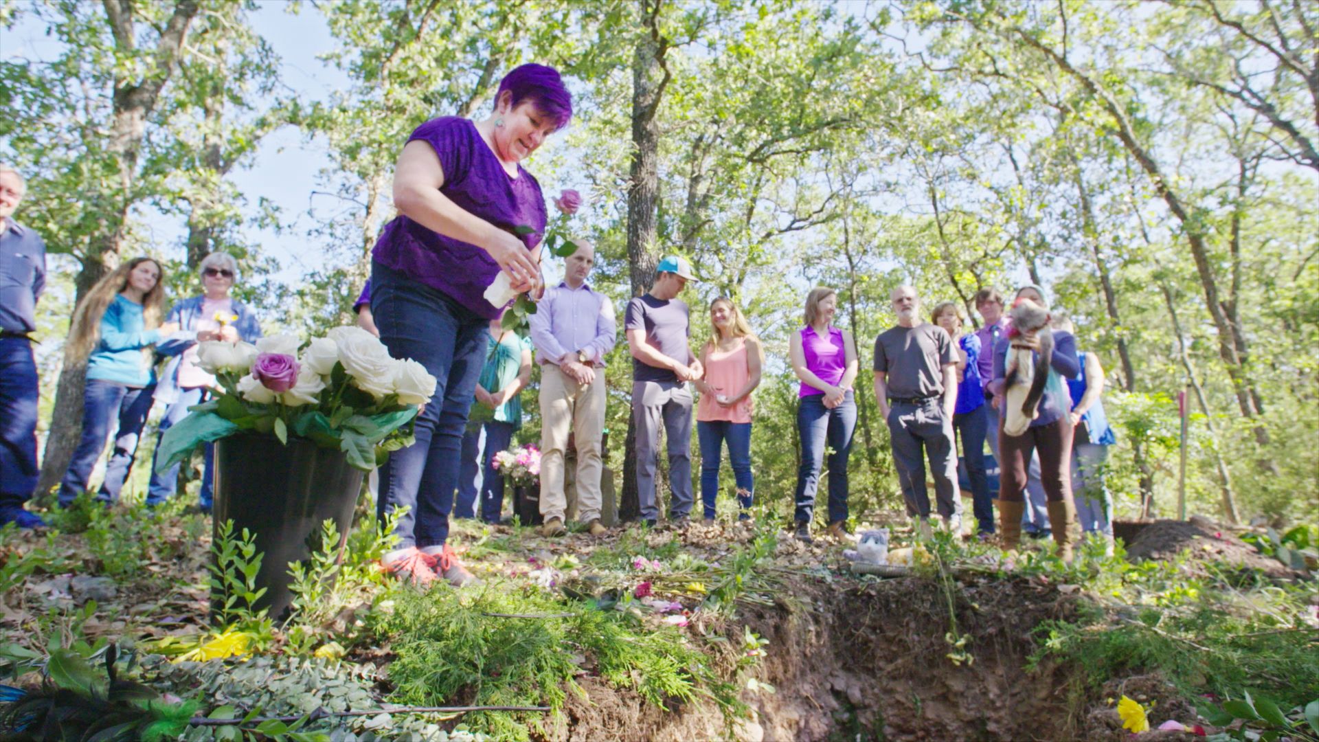 Woman stands over a green burial grave and holds a rose. Several other people stand around the grave.