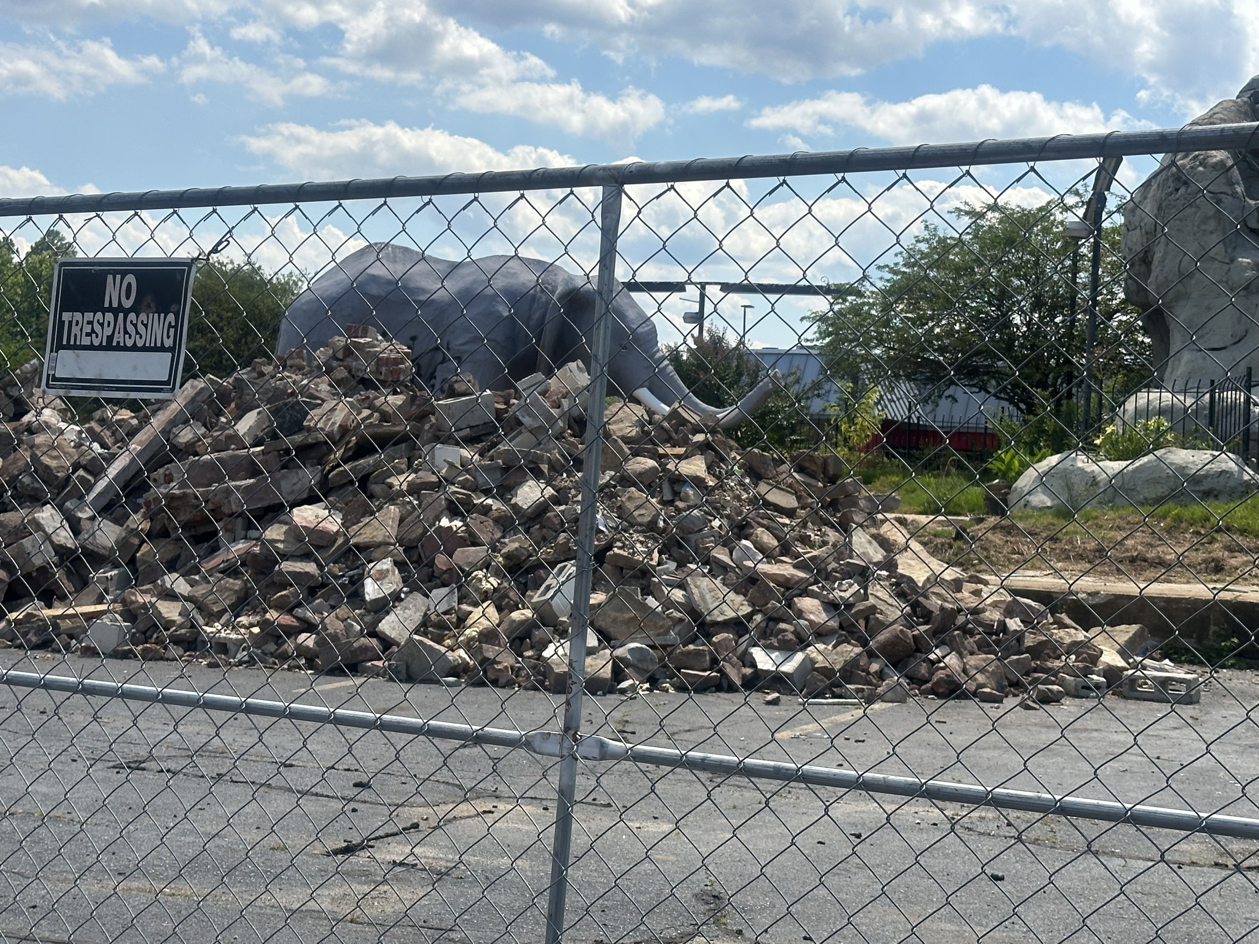 Chain-link fence with a No Trespassing sign blocks view of a rubble-filled construction site; piles of bricks and debris, a tarp-covered mound, large rocks, trees, and a blue sky with clouds.