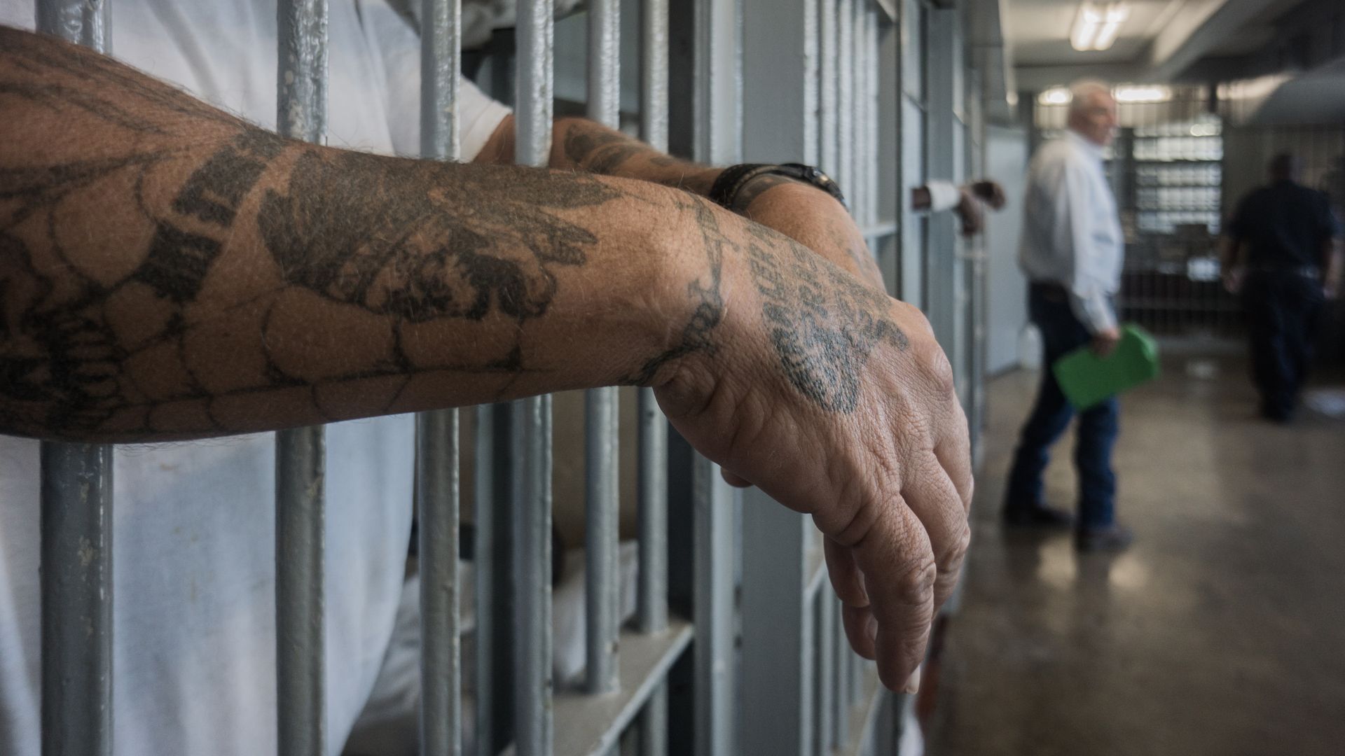 A man's tattooed hands clasp together on the outside of a prison cell.