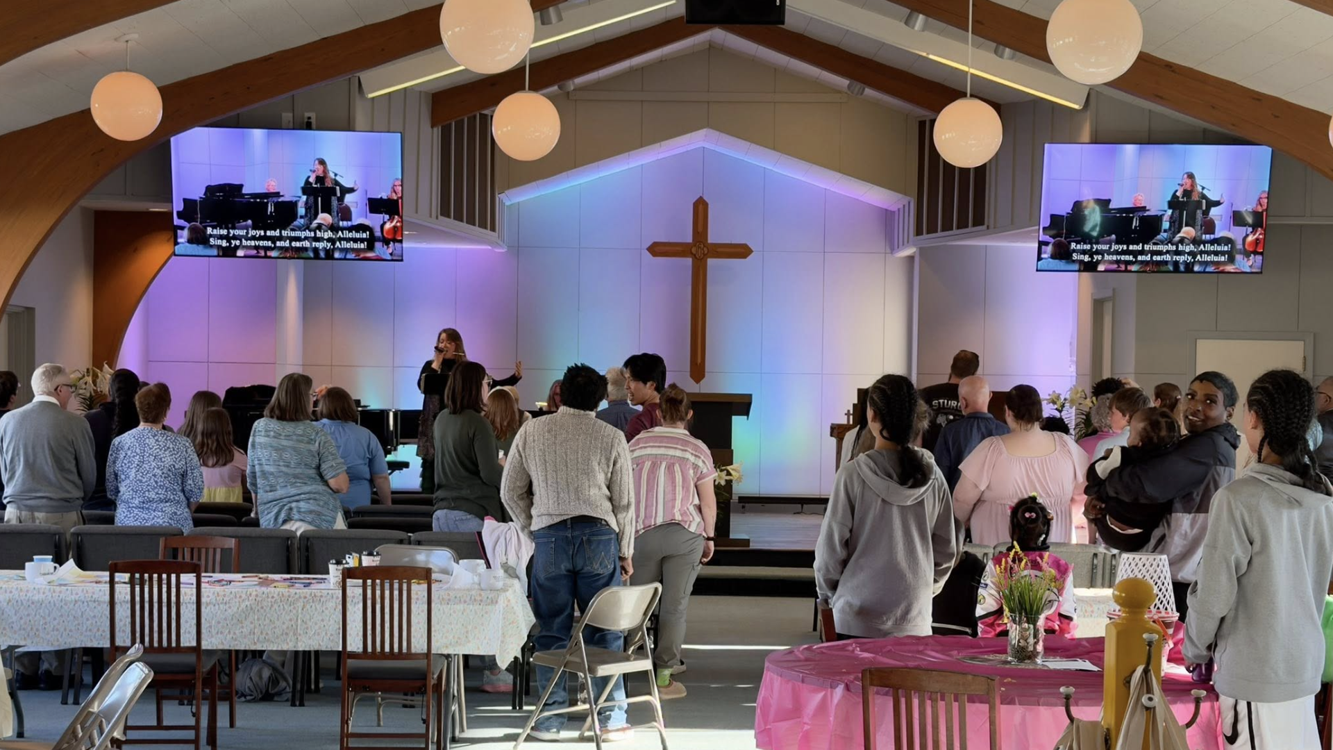 Inside a church, people stand facing a stage with a large wooden cross on a white wall. Colorful uplighting, two screens show a singer with a band; round lamps hang from a vaulted ceiling.