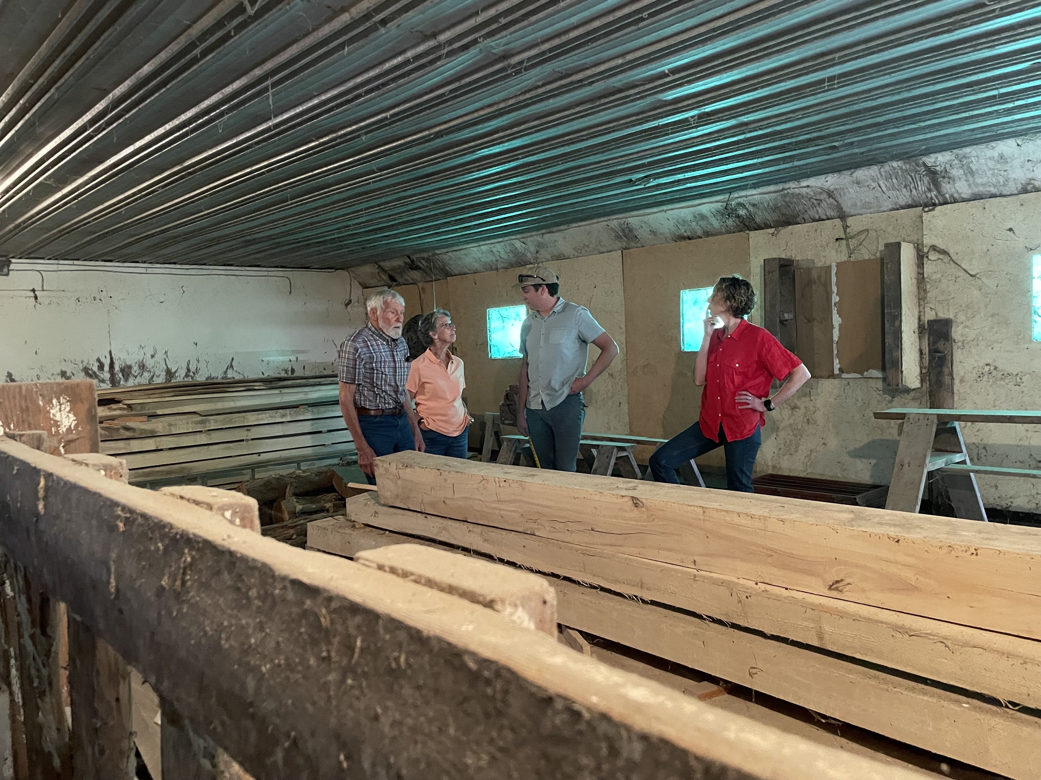 an older man, an older woman, a younger man and a younger woman stand and talk in front of boards of wood in the barn