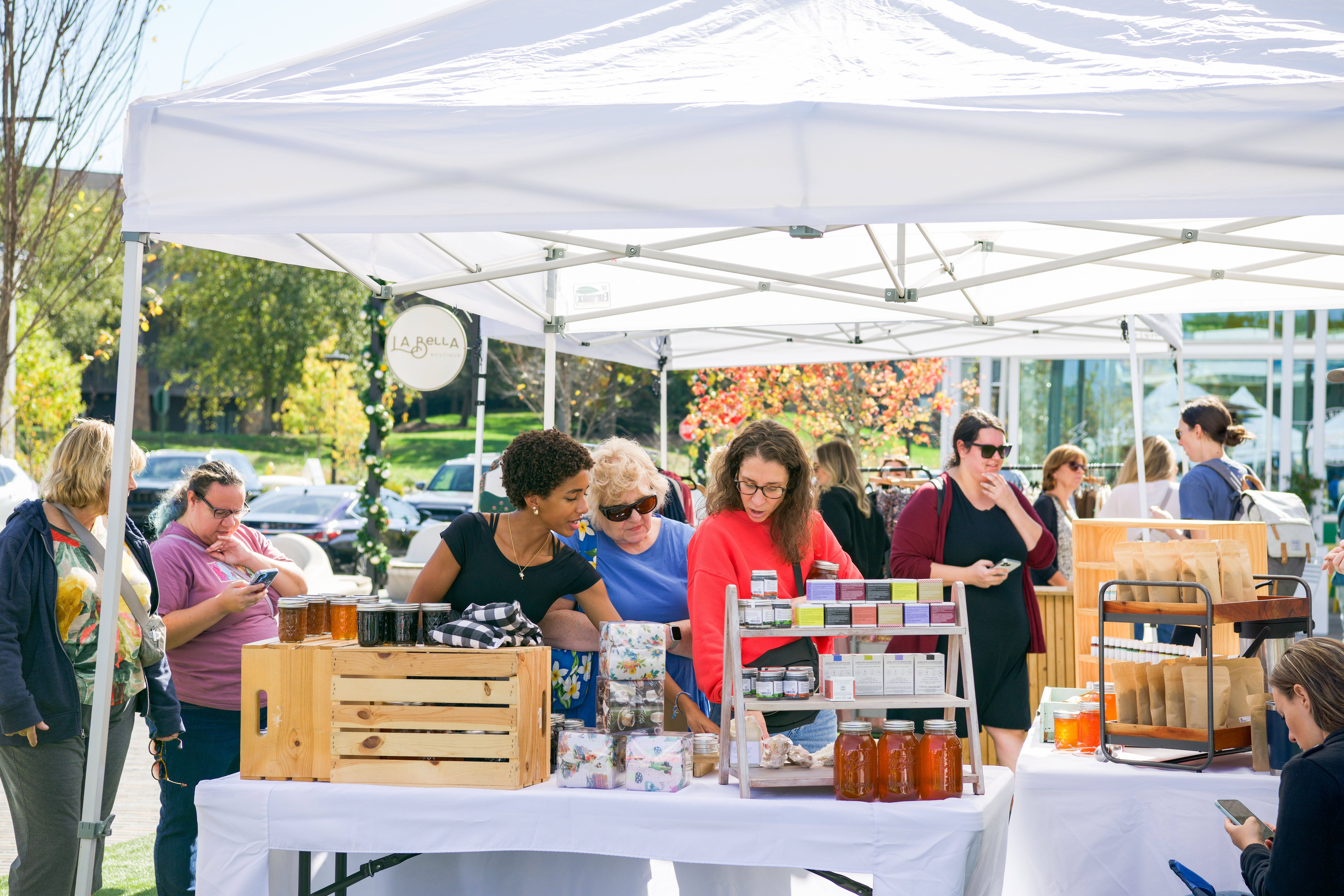 People browse various items including jars, soaps, and packaged goods at an outdoor market under white tents on a sunny day with green trees and autumn foliage in the background.