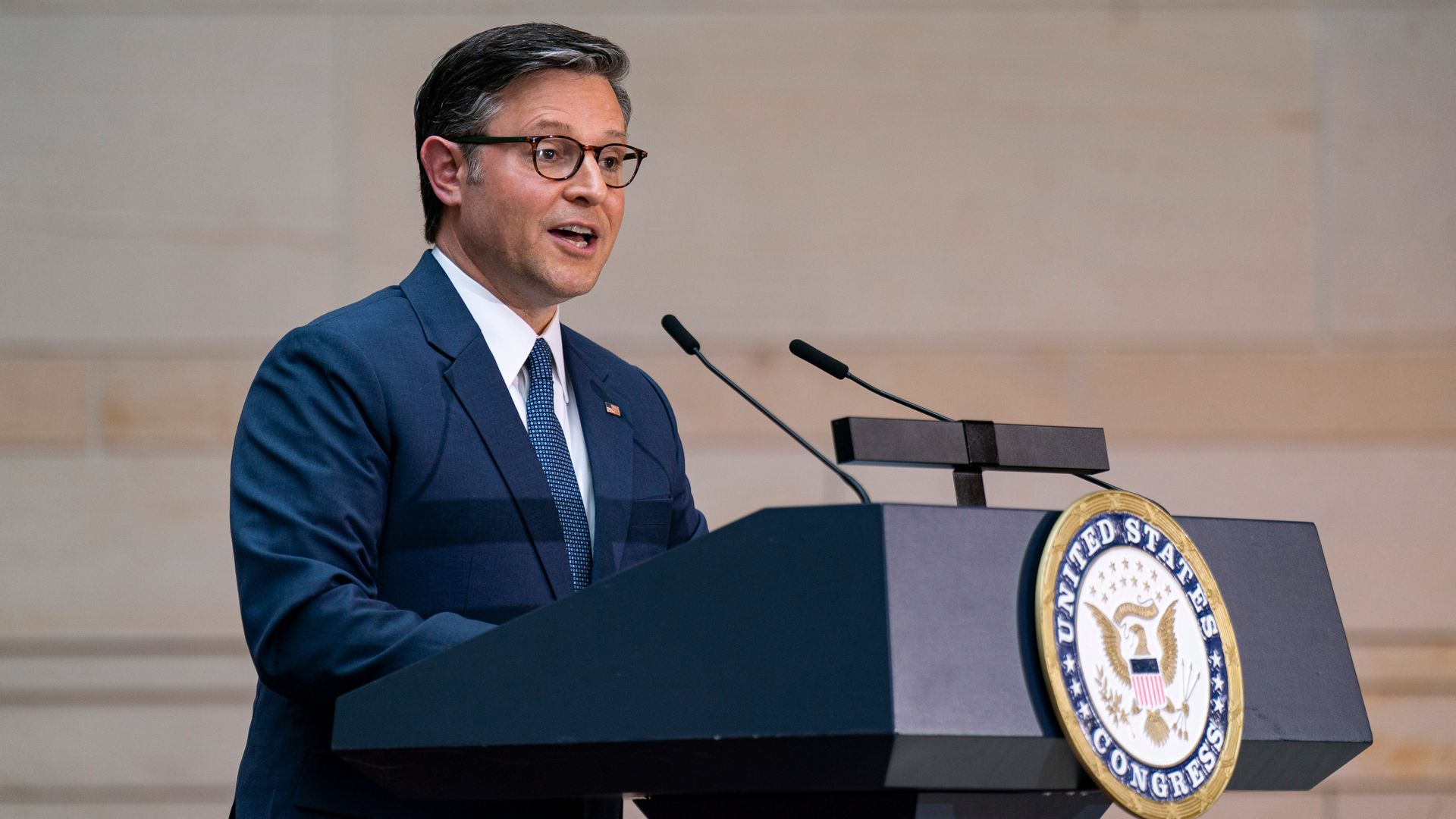 House Speaker Mike Johnson, wearing a blue suit and speaking at a podium in front of a sandstone wall.