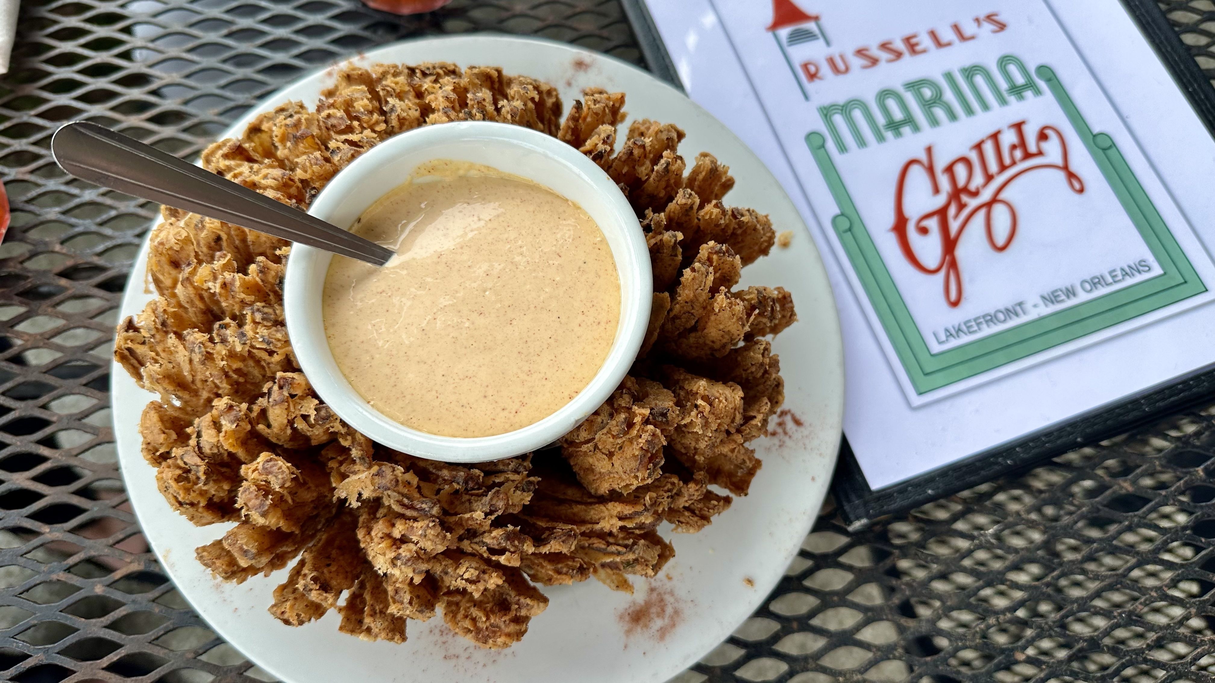 Golden-brown fried onion surround a small white bowl of creamy dipping sauce on a white plate; outdoor metal mesh table with a card reading "RUSSELL'S MARINA" and "NEW ORLEANS" in the background.