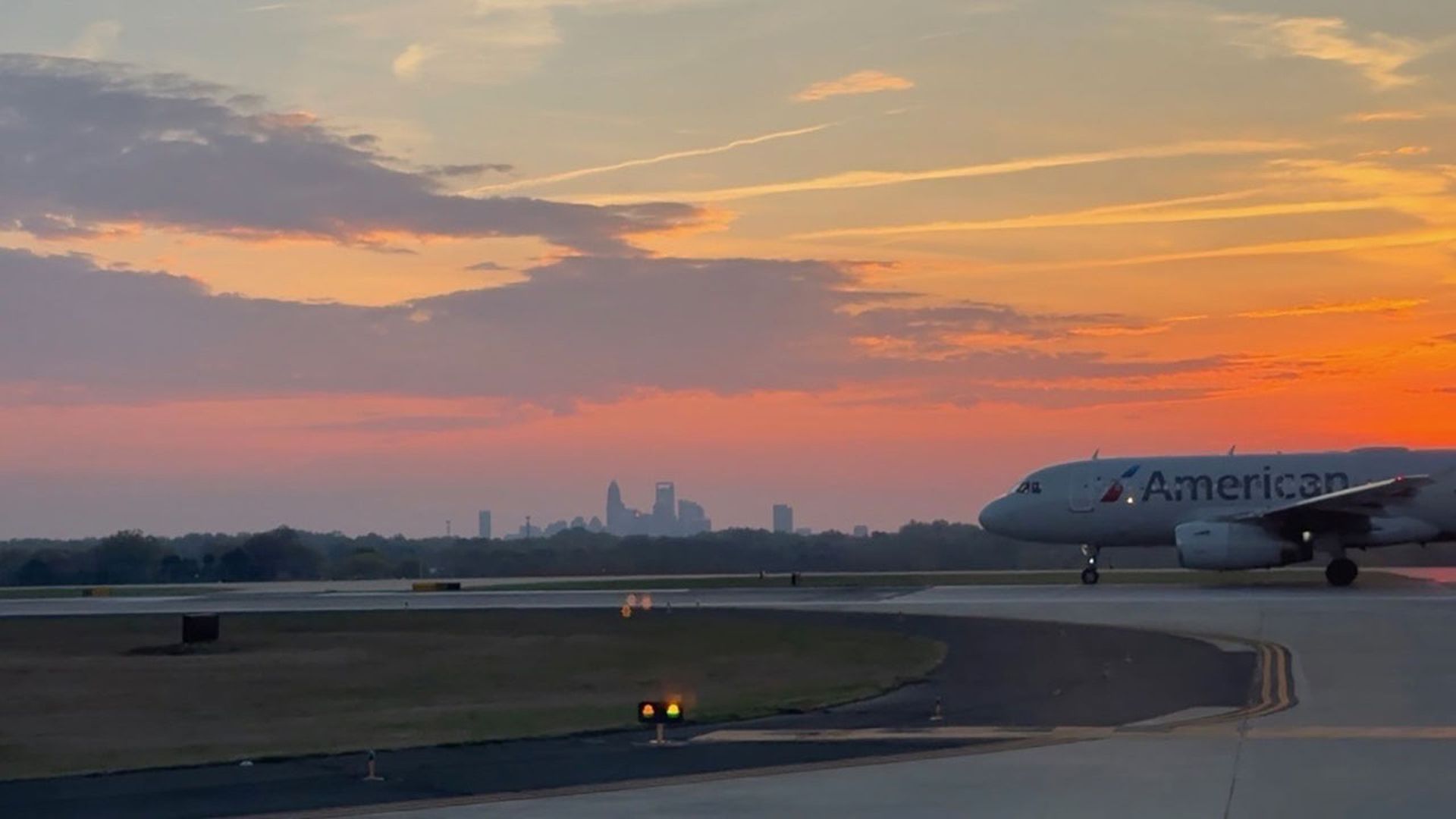 An American Airlines jet taxis on a runway at sunset; orange and pink skies, distant city skyline on the horizon, runway lights and yellow centerlines in view.