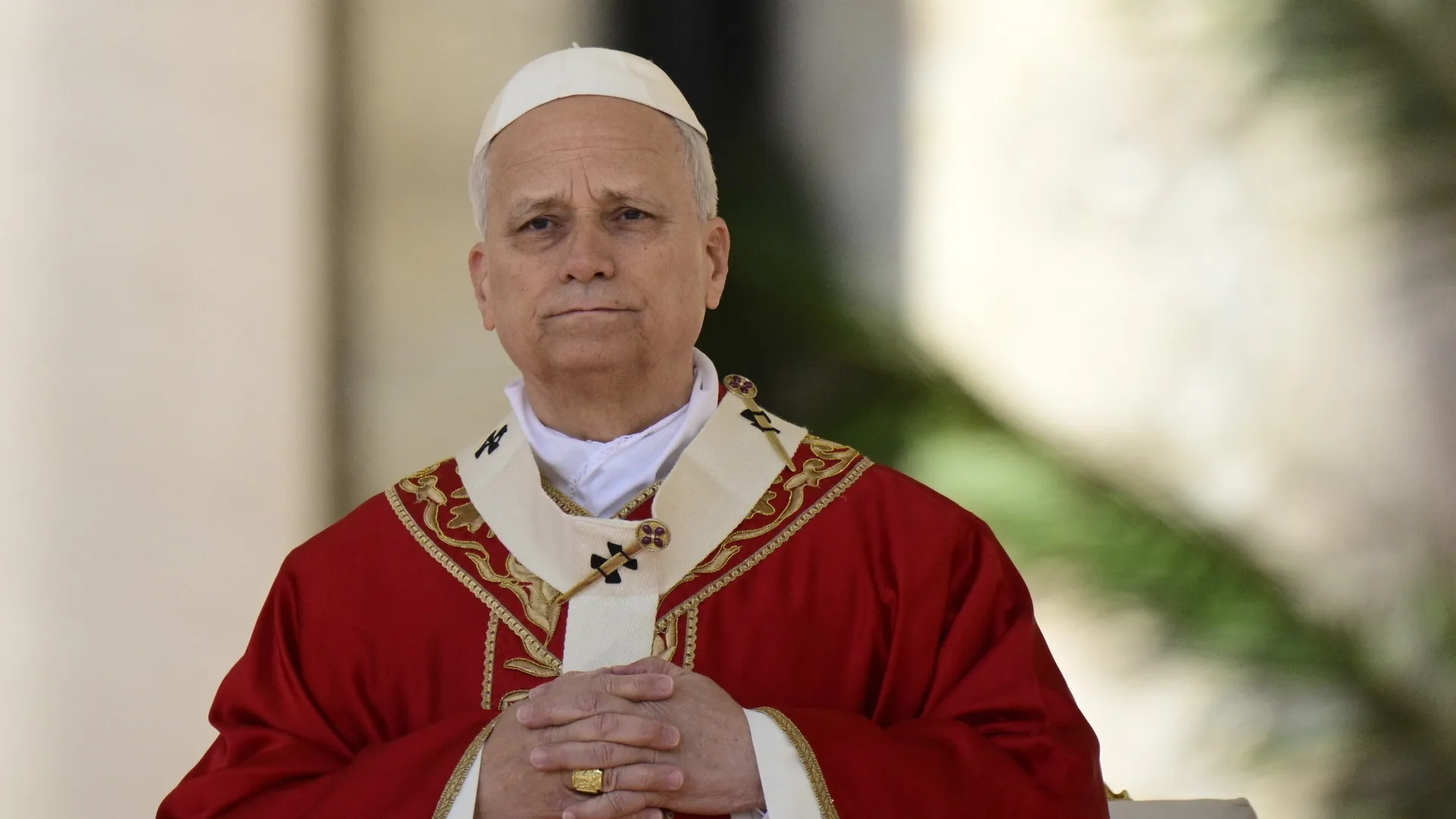 Pope Leo XIV, an older man wearing a white skullcap and ornate red ceremonial robes with gold trim sits with hands folded in front, outdoors with a blurred background.