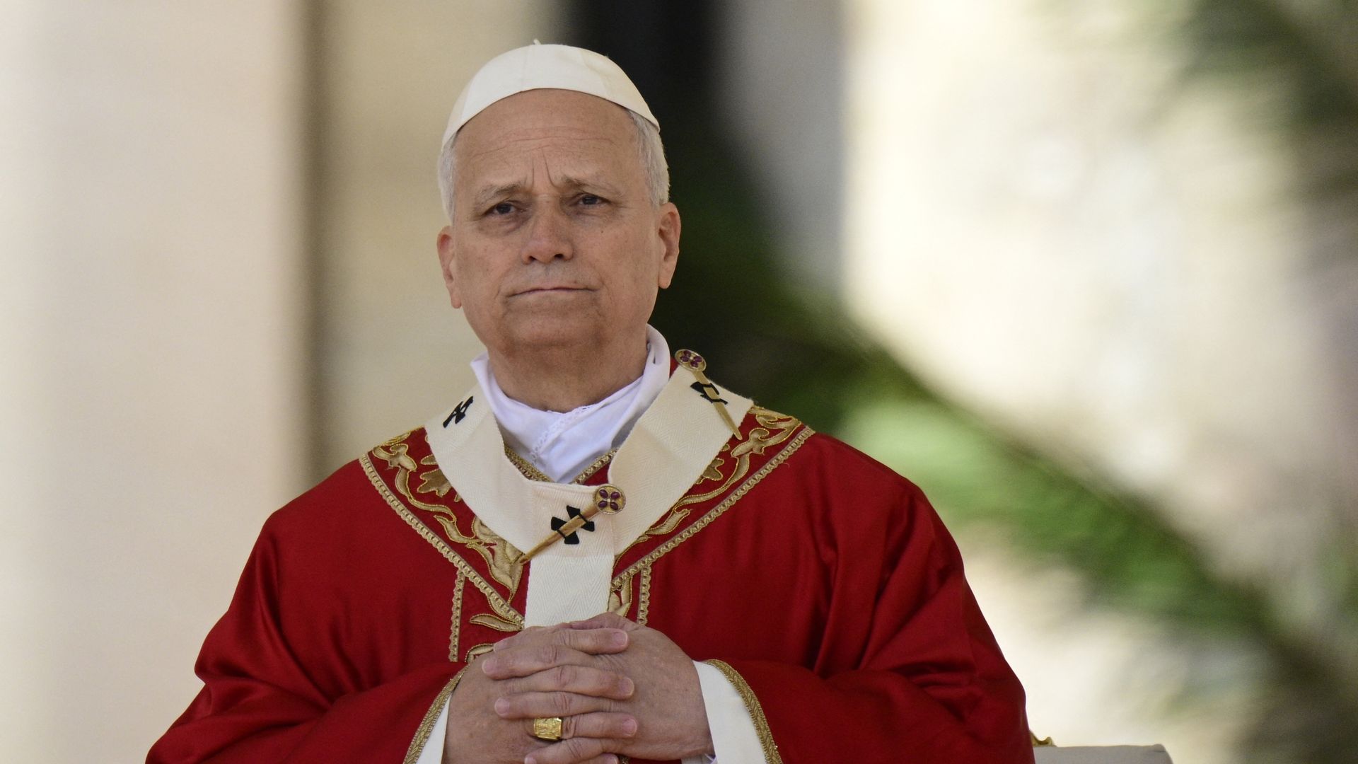 Pope Leo XIV, an older man wearing a white skullcap and ornate red ceremonial robes with gold trim sits with hands folded in front, outdoors with a blurred background.