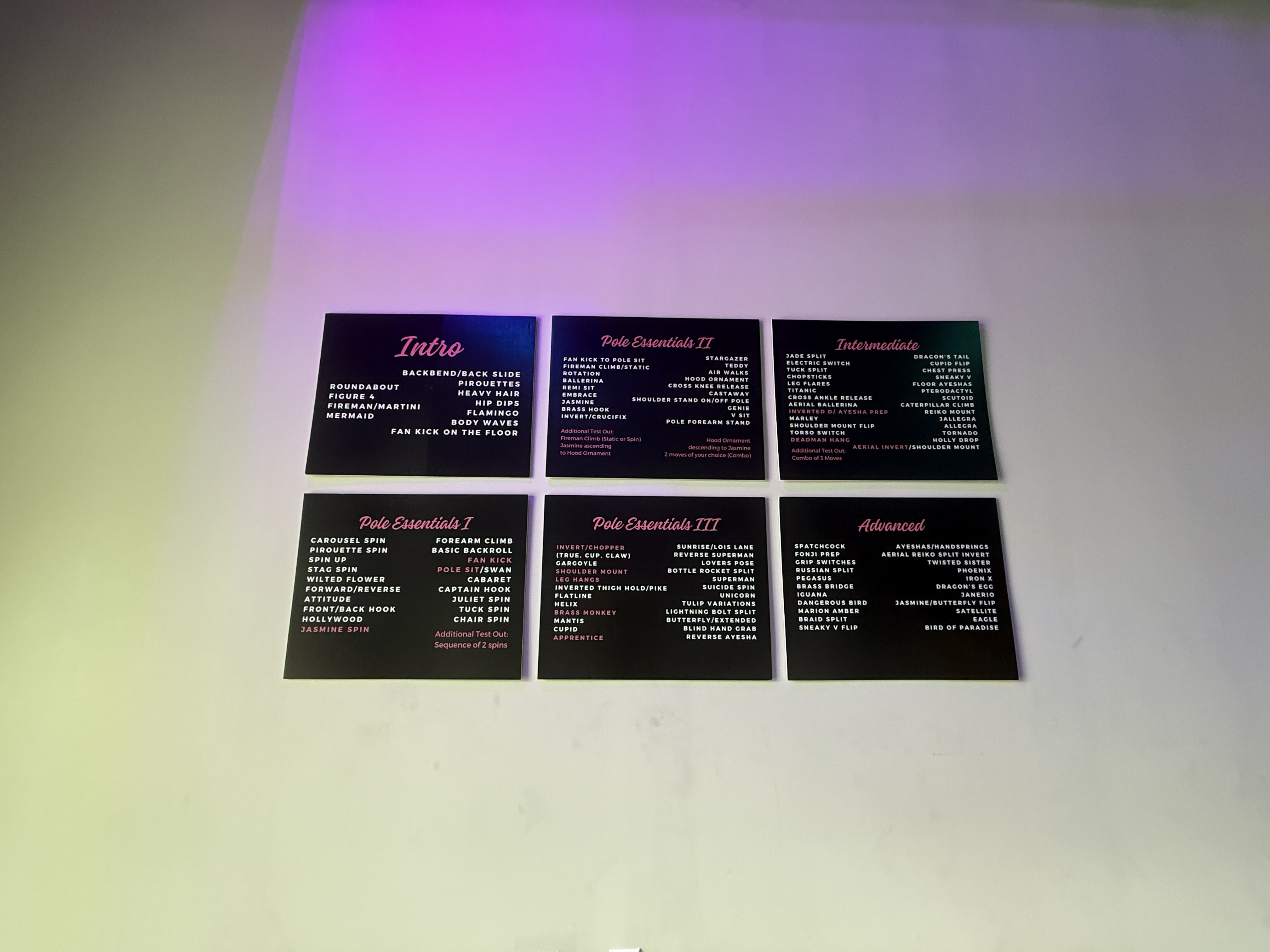 Six black boards with pink and white text listing pole dance moves and skill levels, mounted on a white wall with purple lighting above.