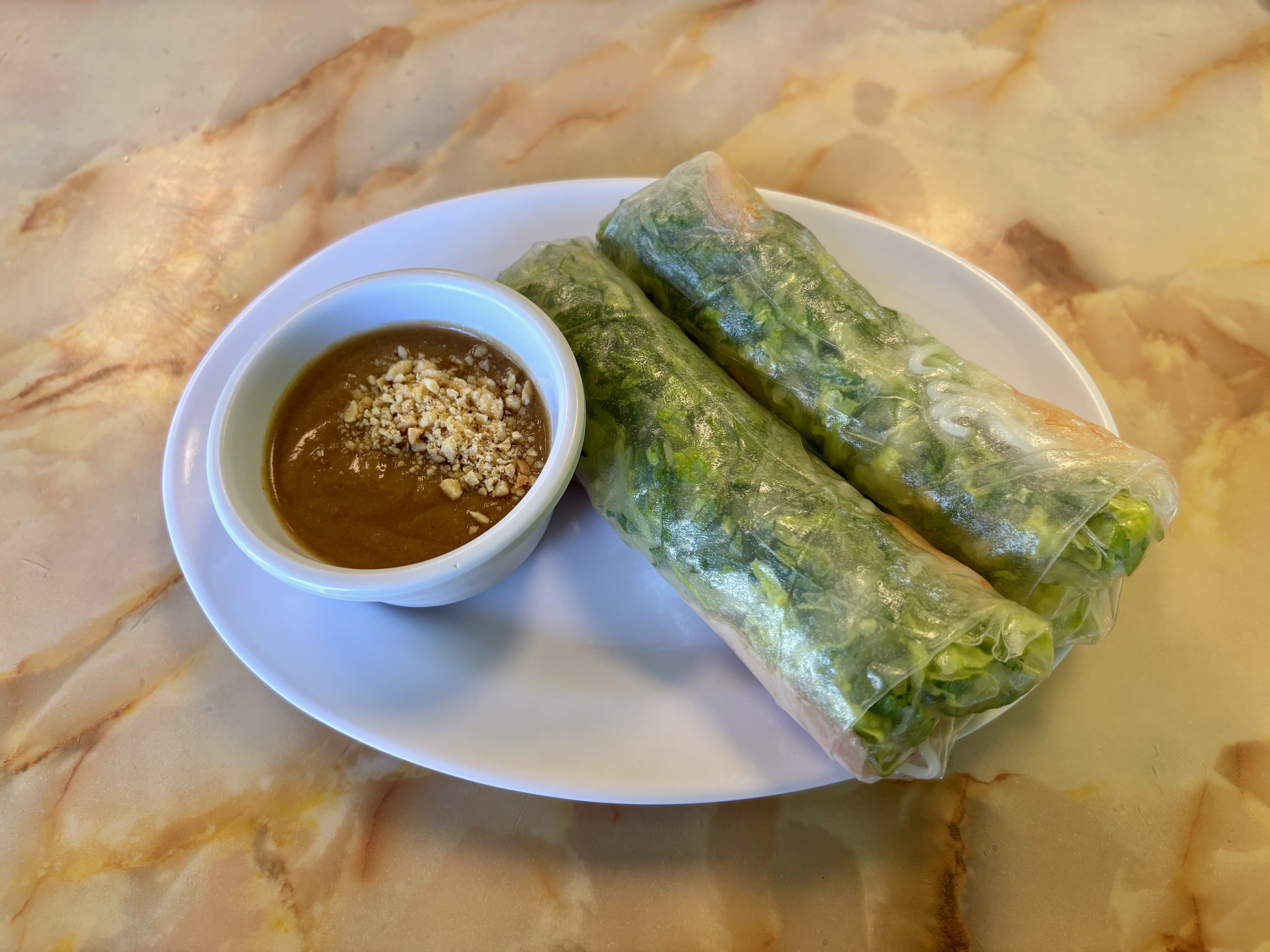 Two Vietnamese spring rolls with green leafy vegetables wrapped in translucent rice paper, served on a white plate with a small bowl of brown peanut sauce topped with crushed peanuts, on a marble table.