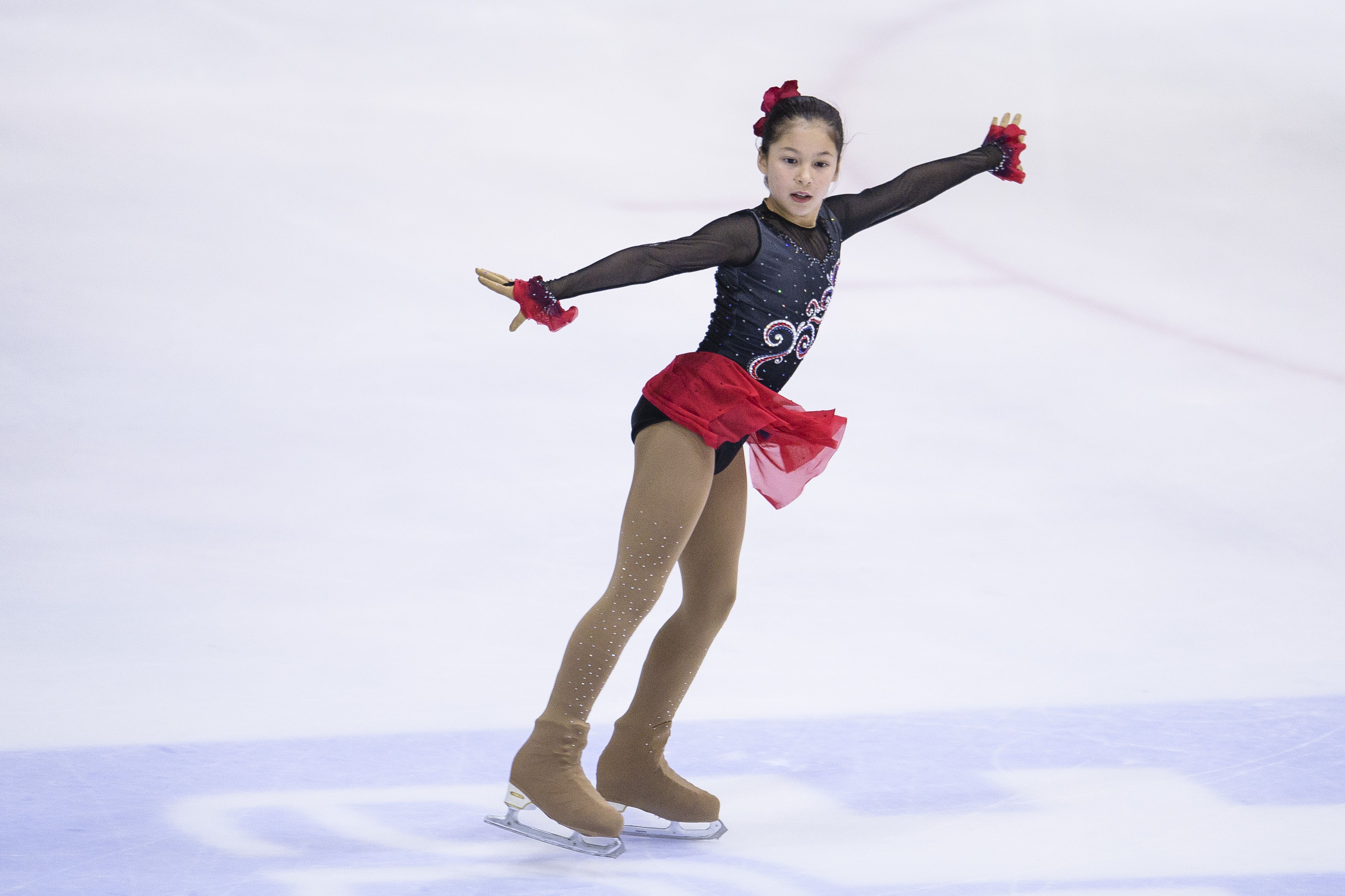 Young female figure skater in a black and red costume with long sleeves and beige tights mid-performance on ice rink, arms extended for balance.