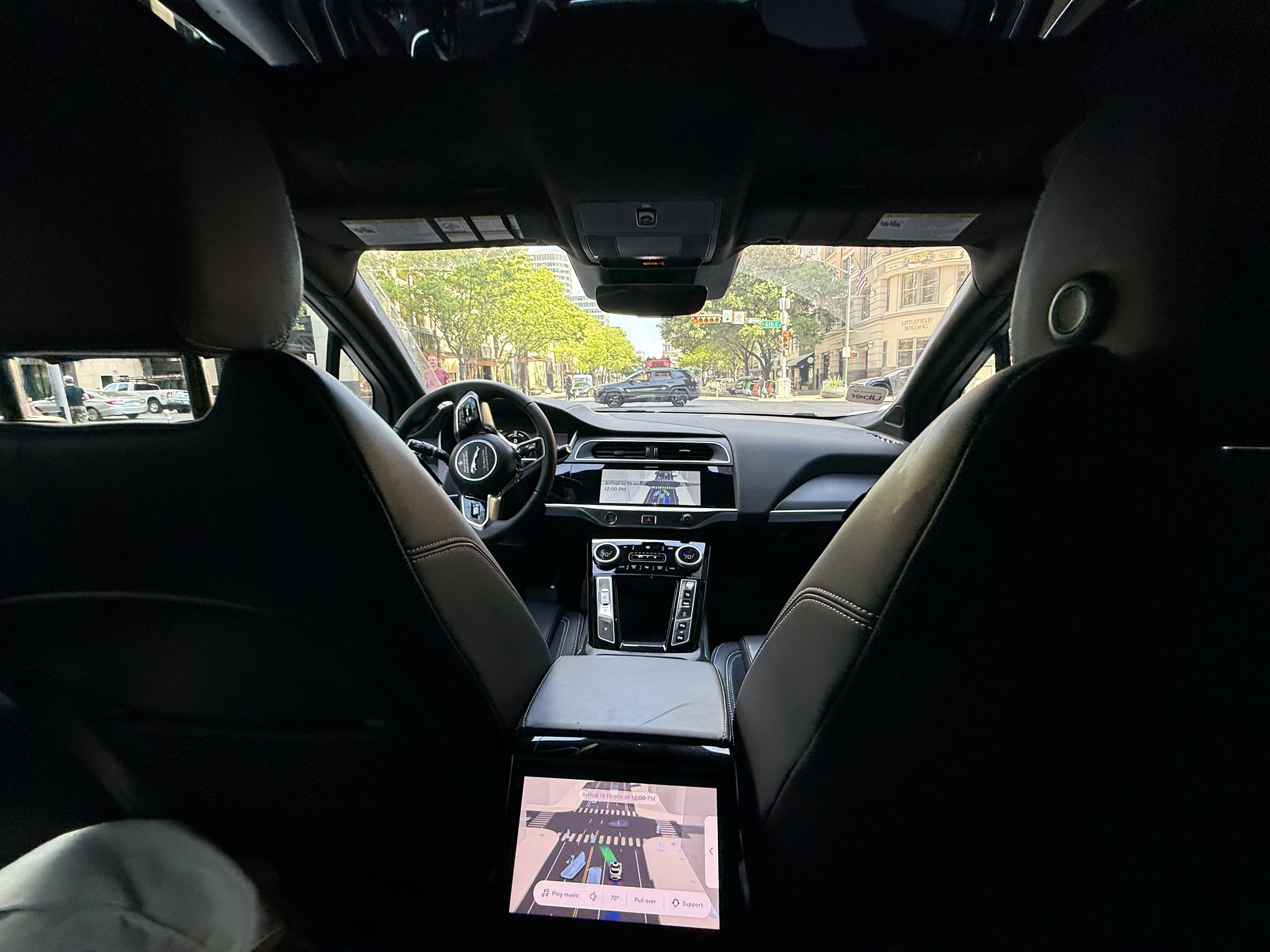 Interior of a dark luxury car photographed from the back seat; front seats and steering wheel visible, center console screen and gear area. Through the windshield, a tree-lined city street.