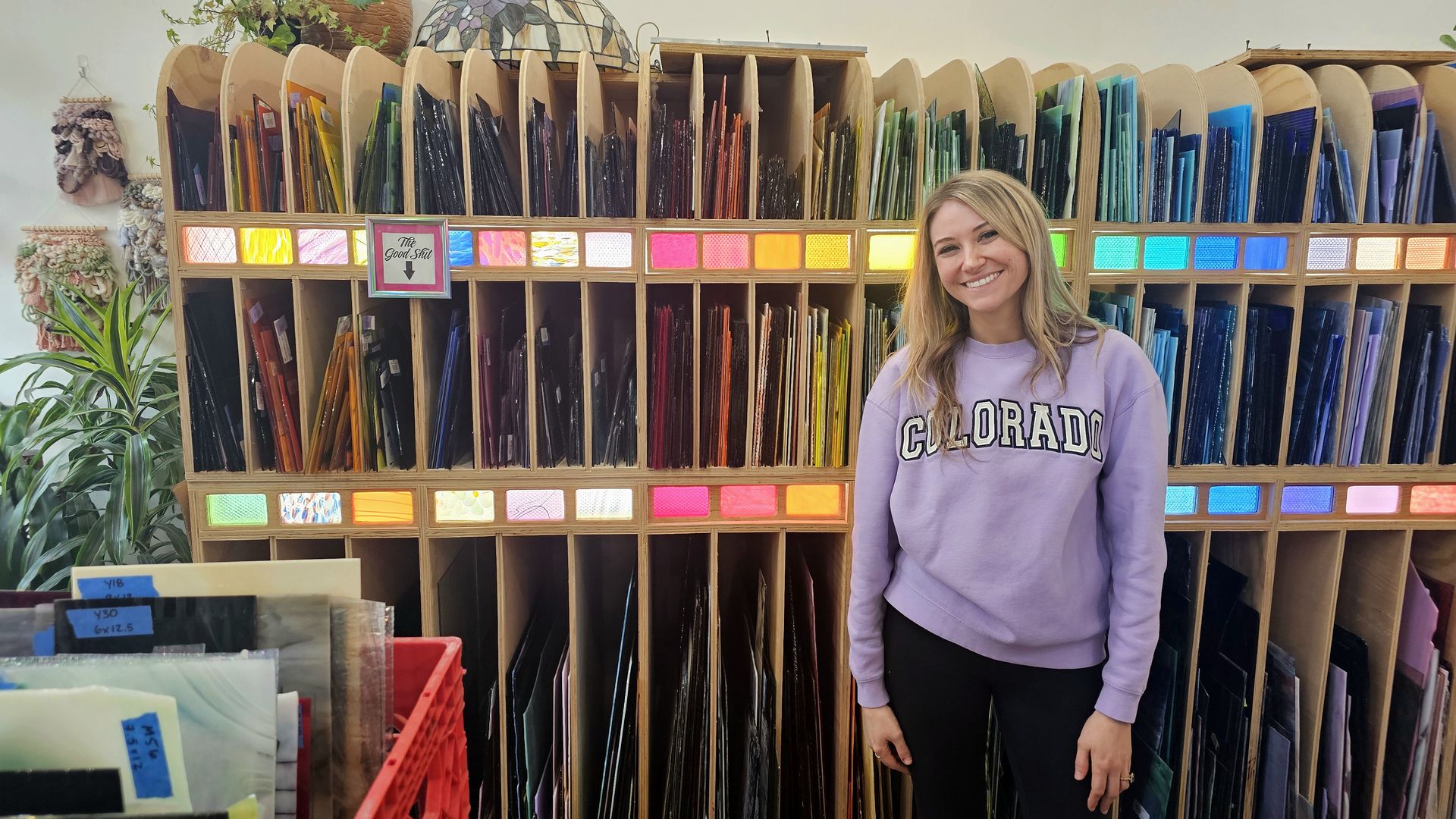 Smiling woman with light hair in a lavender Colorado sweatshirt stands beside a rainbow display of colorful glass sheets organized by hue in wooden cubbies, with plants visible on the left.