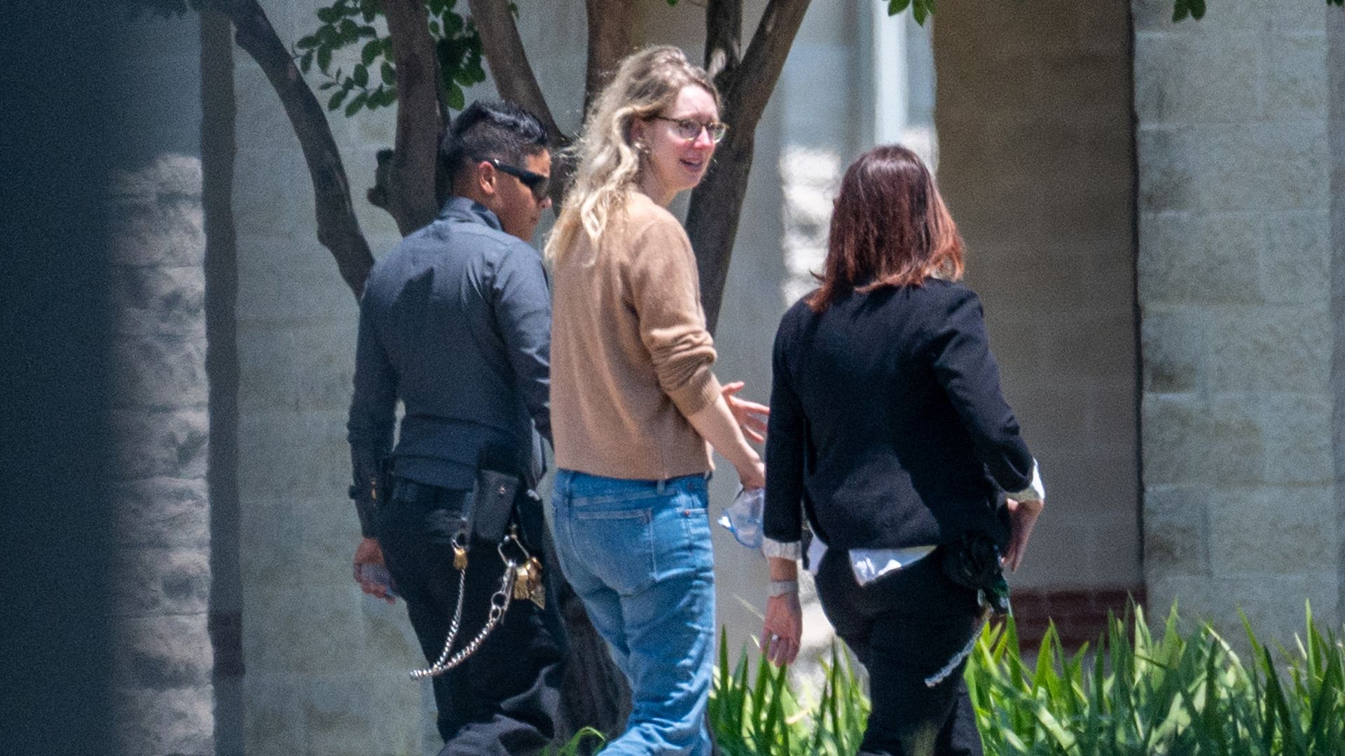 Three people walk along a sidewalk by a stone building with trees; a man in dark clothes and sunglasses, a blonde woman in beige sweater and jeans, and another person; yellow tape along the grass.