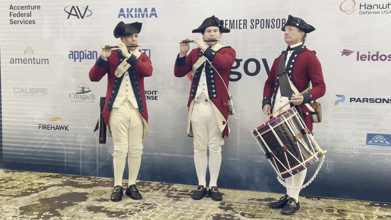 Three men in red 18th-century coats and tricorne hats stand on a stage: two flutists and a drummer with a large drum, backdrop covered in sponsor logos.