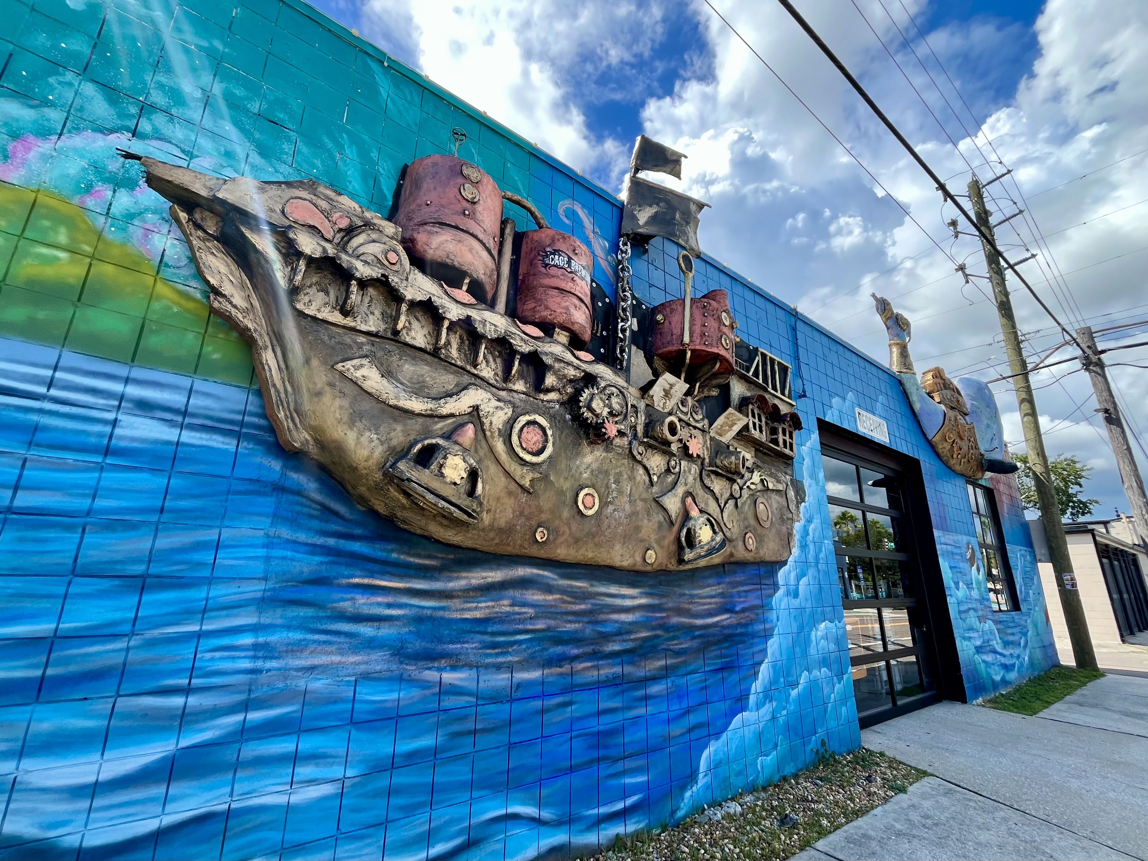 A colorful mural on a blue tiled wall shows a large rust-colored mechanical sculpture with chains and rock-like shapes, under a bright cloudy sky and overhead power lines.