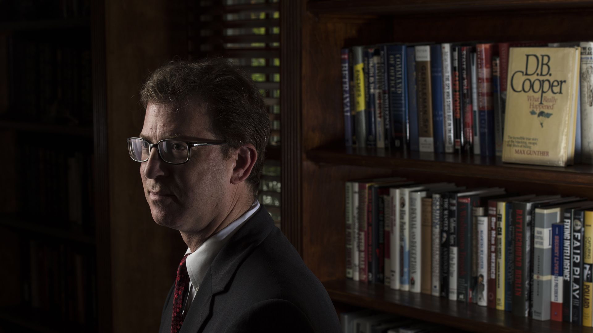 Mark Zaid — wearing a black suit, a collared shirt, a red tie and glasses — looks on while standing in front of bookshelves in a dimly lit room.