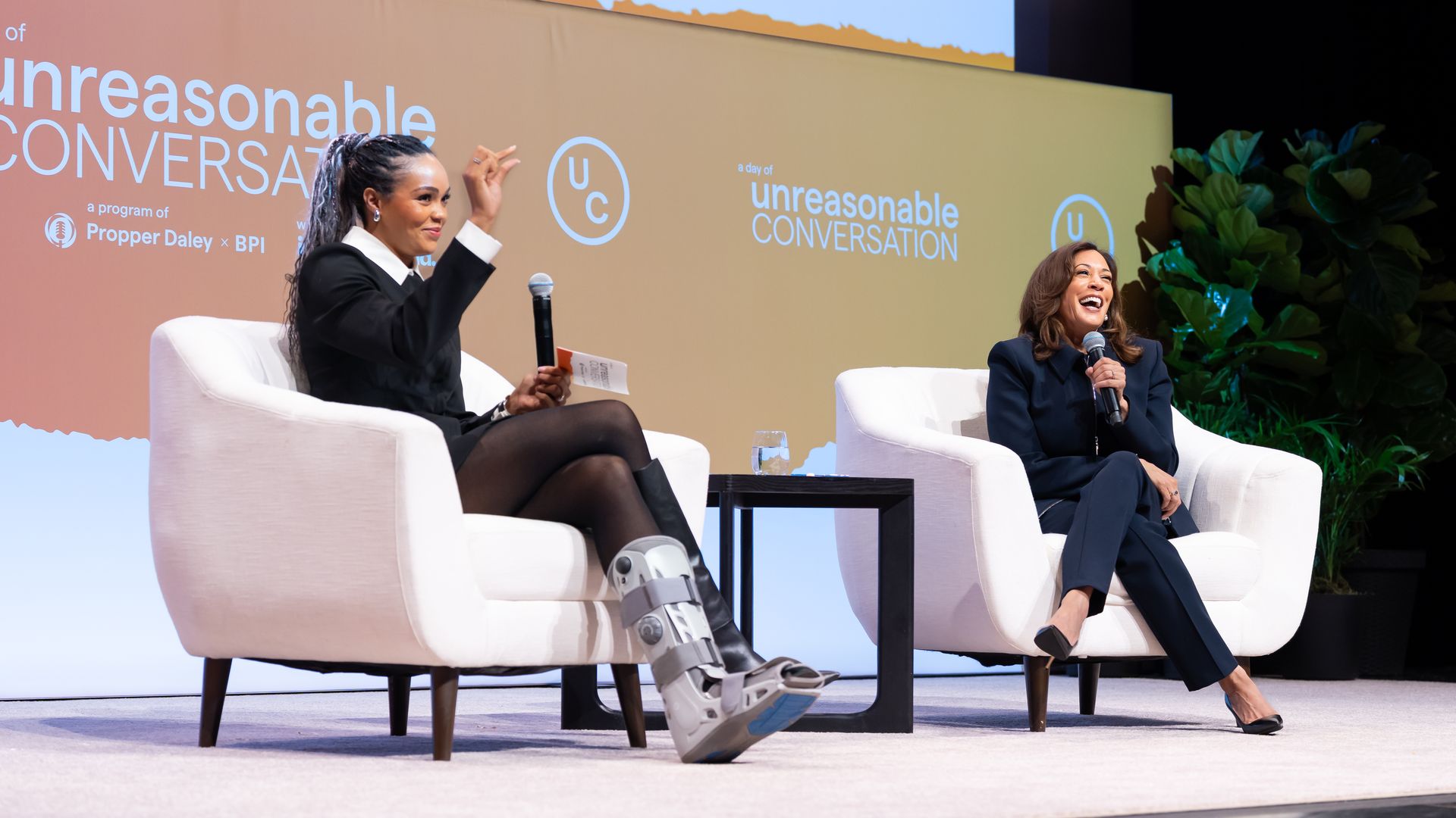 Two women are seated on white armchairs on a stage during "a day of unreasonable conversation" event; one woman wears a black outfit with a leg brace, the other in a dark suit, both holding microphones.