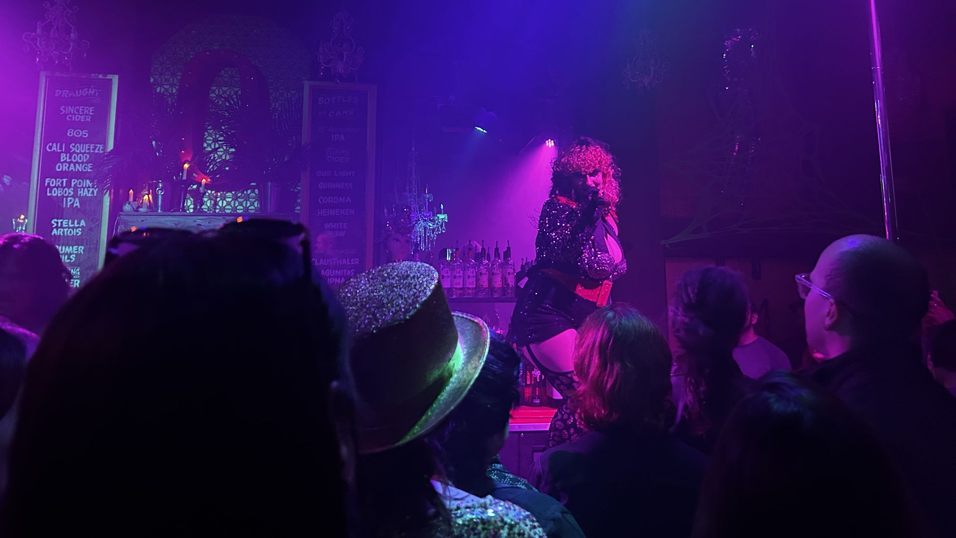 A performer in a glittery black outfit and curly hair sings on a lit stage at a bar with chandeliers, colorful purple and blue lighting, and a crowd watching.