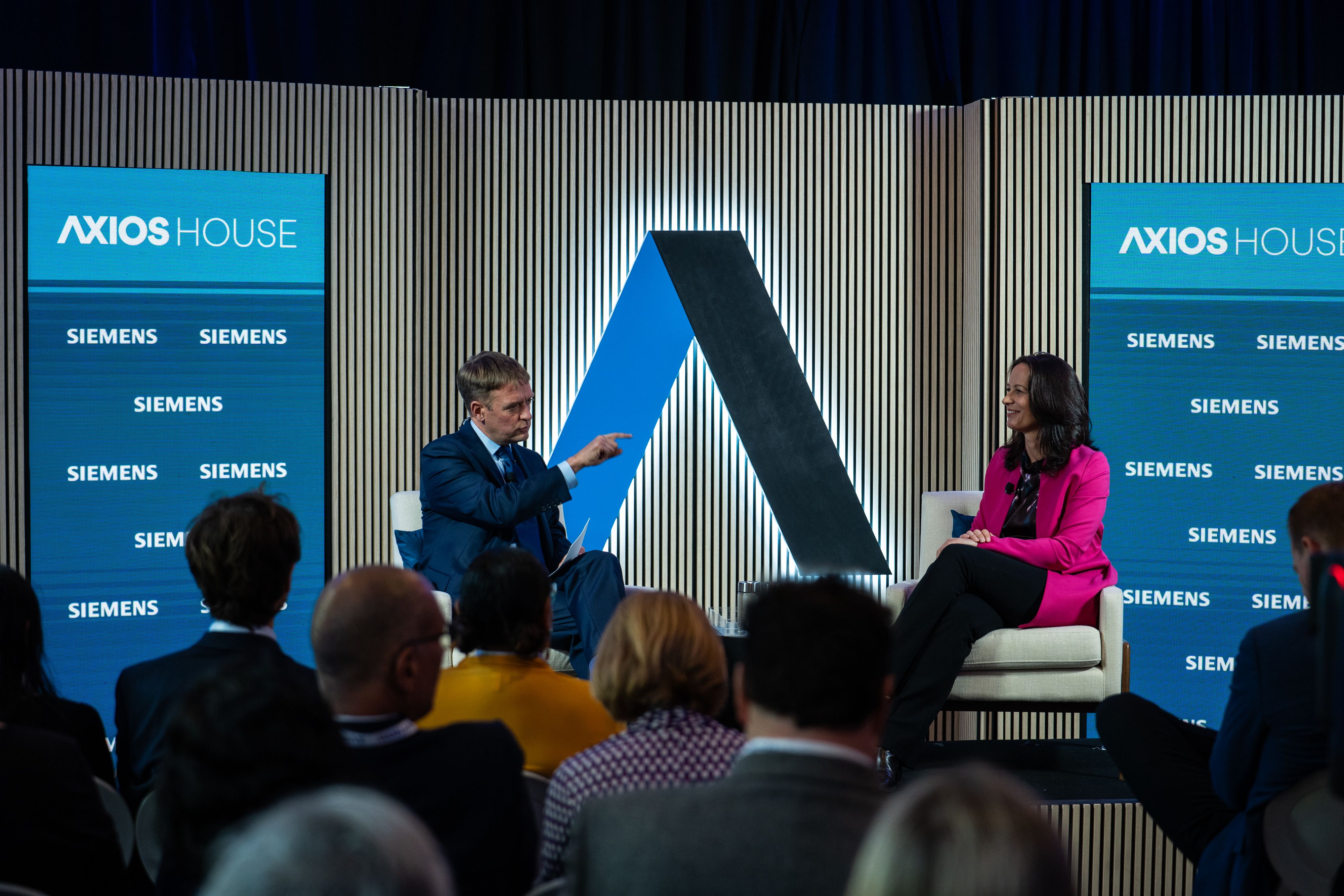 Panel discussion on a stage with Axios House banners and Siemens logos. A man in a blue suit gestures toward a woman in pink; a large blue and black angular sculpture sits between them, audience in front.