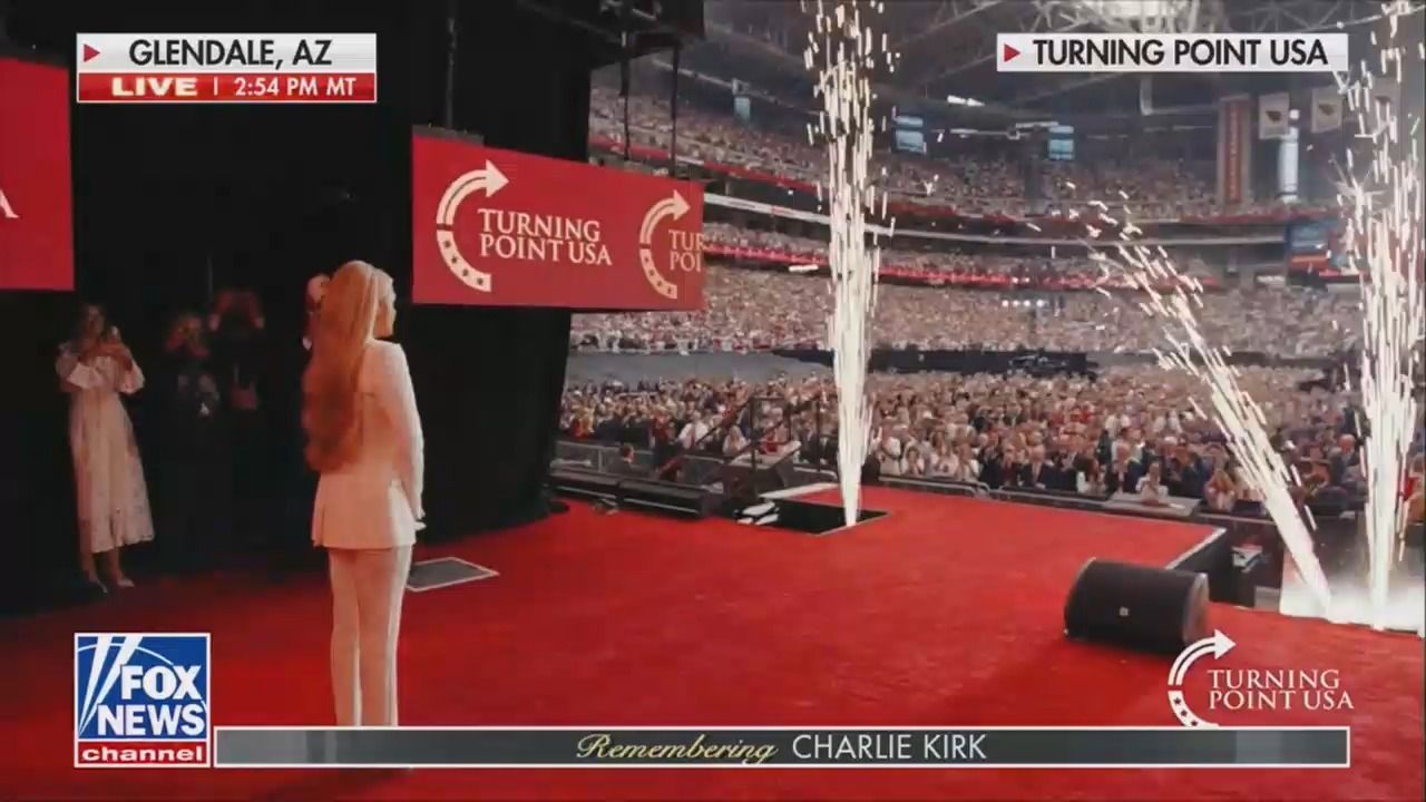 Erika Kirk stands on a red stage at a memorial service for her husban Charlie Kirk. Fireworks shoot up near the stage. Signs say "Turning Point USA." Broadcast live from Glendale, AZ on Fox News.