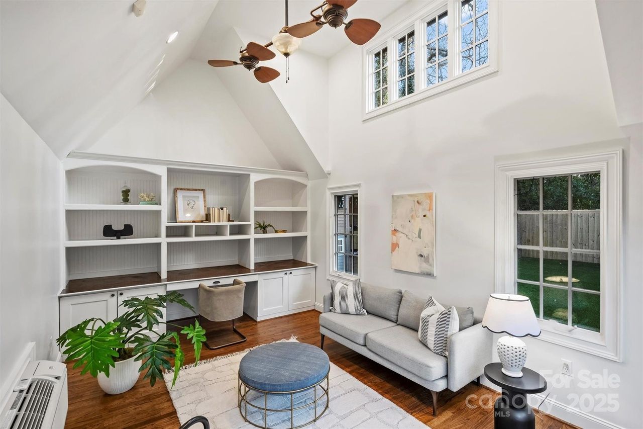 Bright living room with high ceilings, white walls, built-in shelves and desk, gray sofa with patterned pillows, blue round ottoman, white rug, green plant, and ceiling fans with wooden blades.