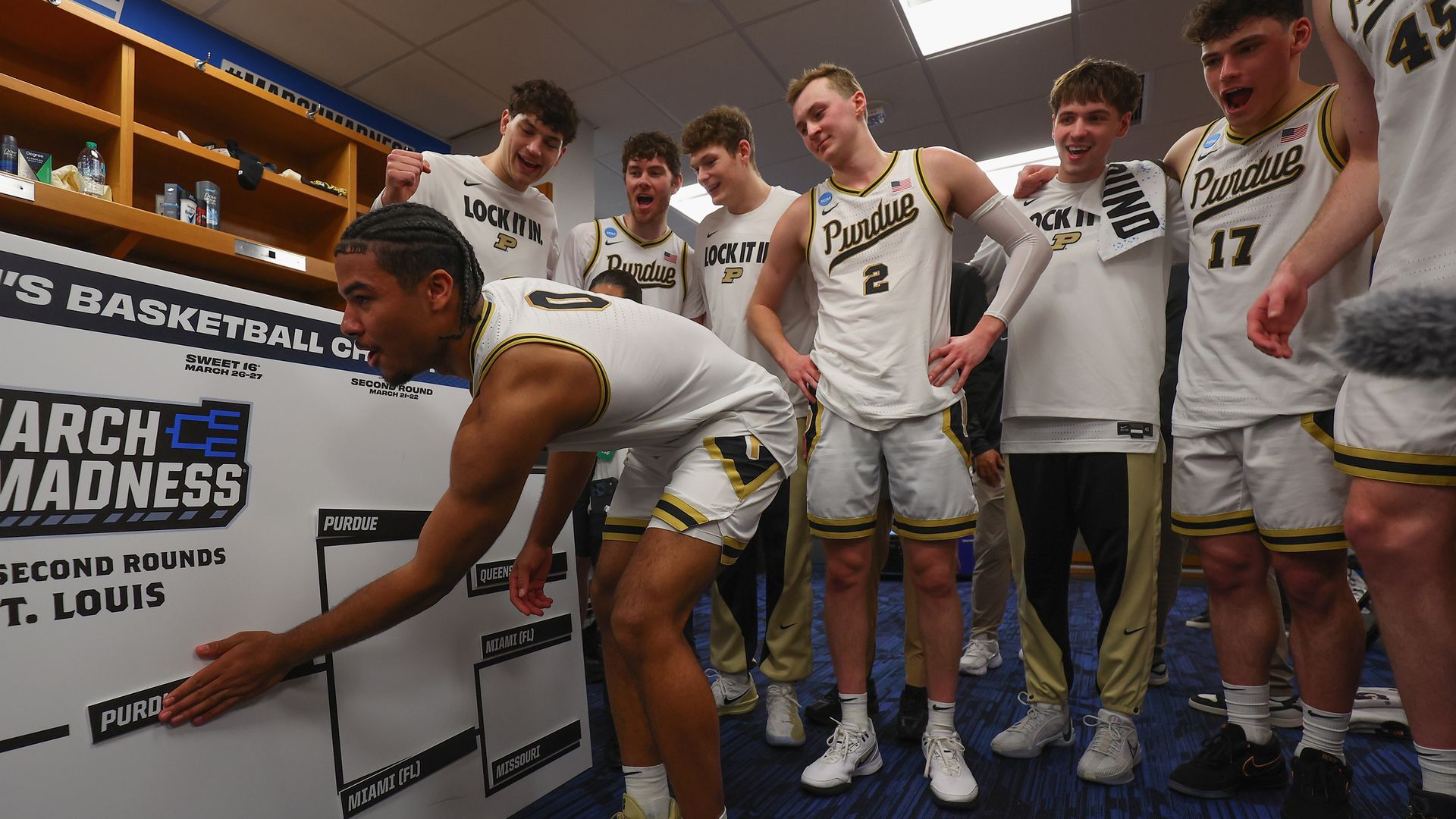 Purdue basketball players in white and gold uniforms surround a locker-room board labeled "March Madness" as one player leans forward, pointing at the bracket while teammates cheer.
