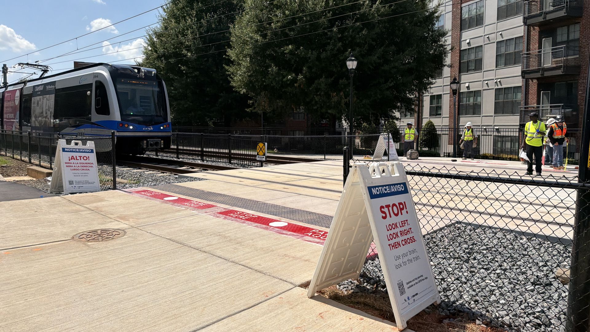 Blue and white tram on tracks near a construction site; workers in high-visibility vests stand by a chain-link fence beside a red brick apartment building, with a large tree and blue sky.