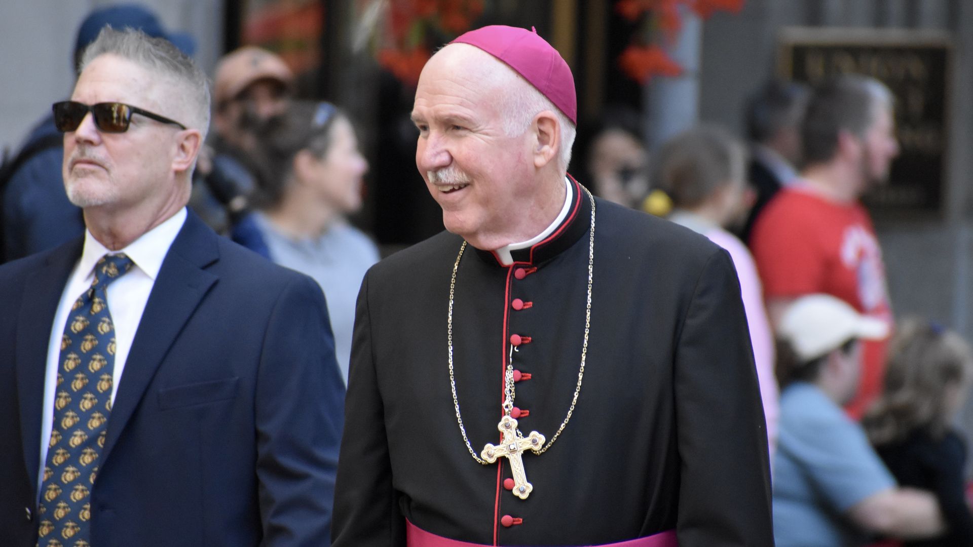 A man in a black clerical robe with red trim and a pink sash wears a pink skullcap and a large cross necklace, smiling. Another man in a blue suit and sunglasses walks beside him.
