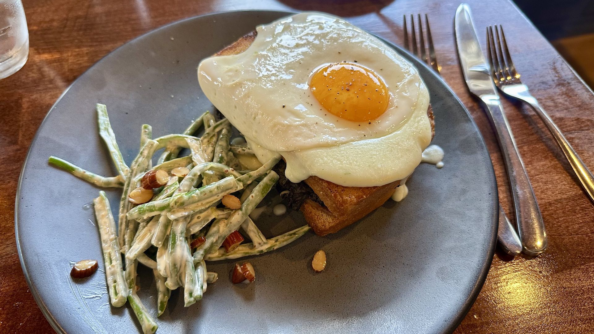 A gray plate on a wooden table features a sunny-side-up egg on toast with creamy sauce, paired with a green bean salad tossed with almonds; a glass of water and cutlery are nearby.