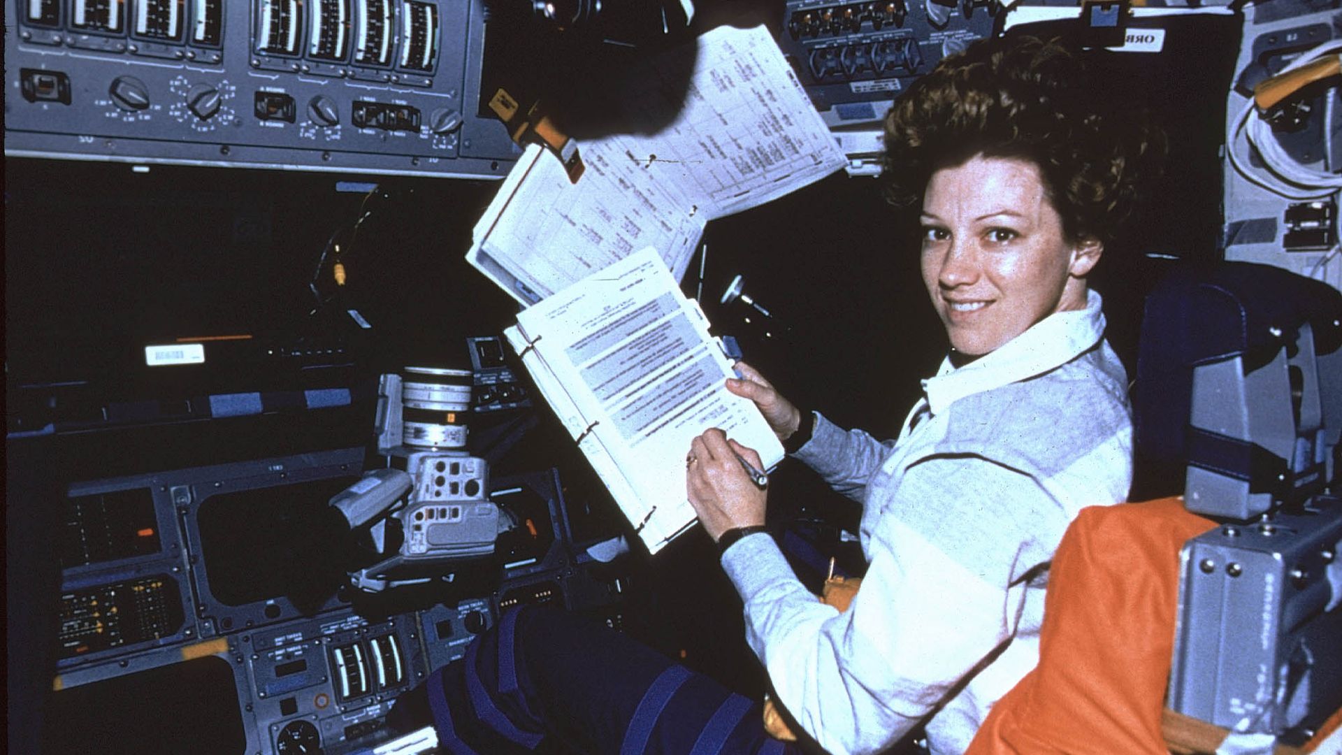 Eileen Collins in a spacecraft cockpit, smiling, holding and reading a binder of documents amid control panels, switches, and equipment.