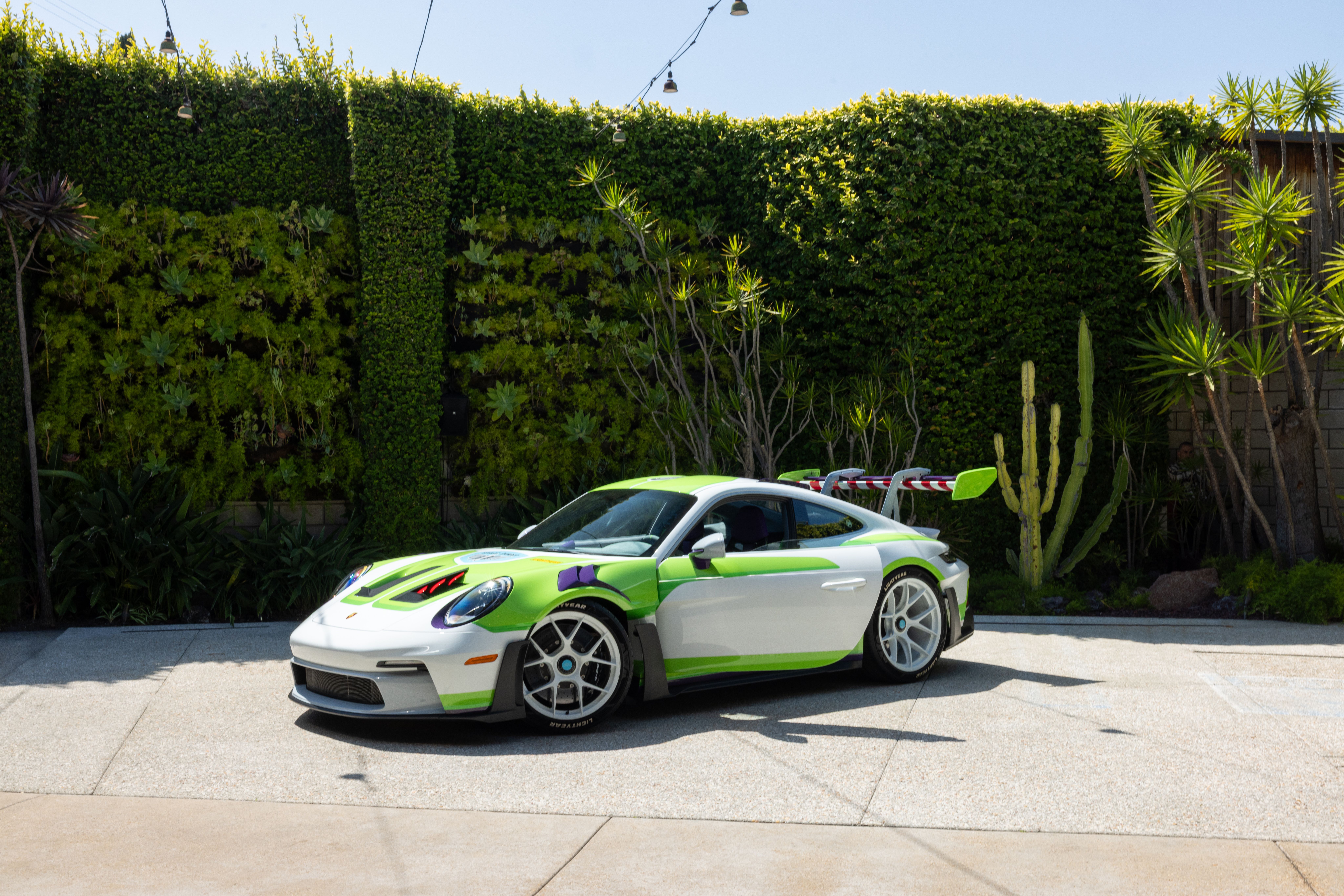 Bright green and white sports car with purple accents parked on light pavement, in front of a tall hedged wall with string lights overhead; right side shows desert plants and cacti under a clear blue sky.