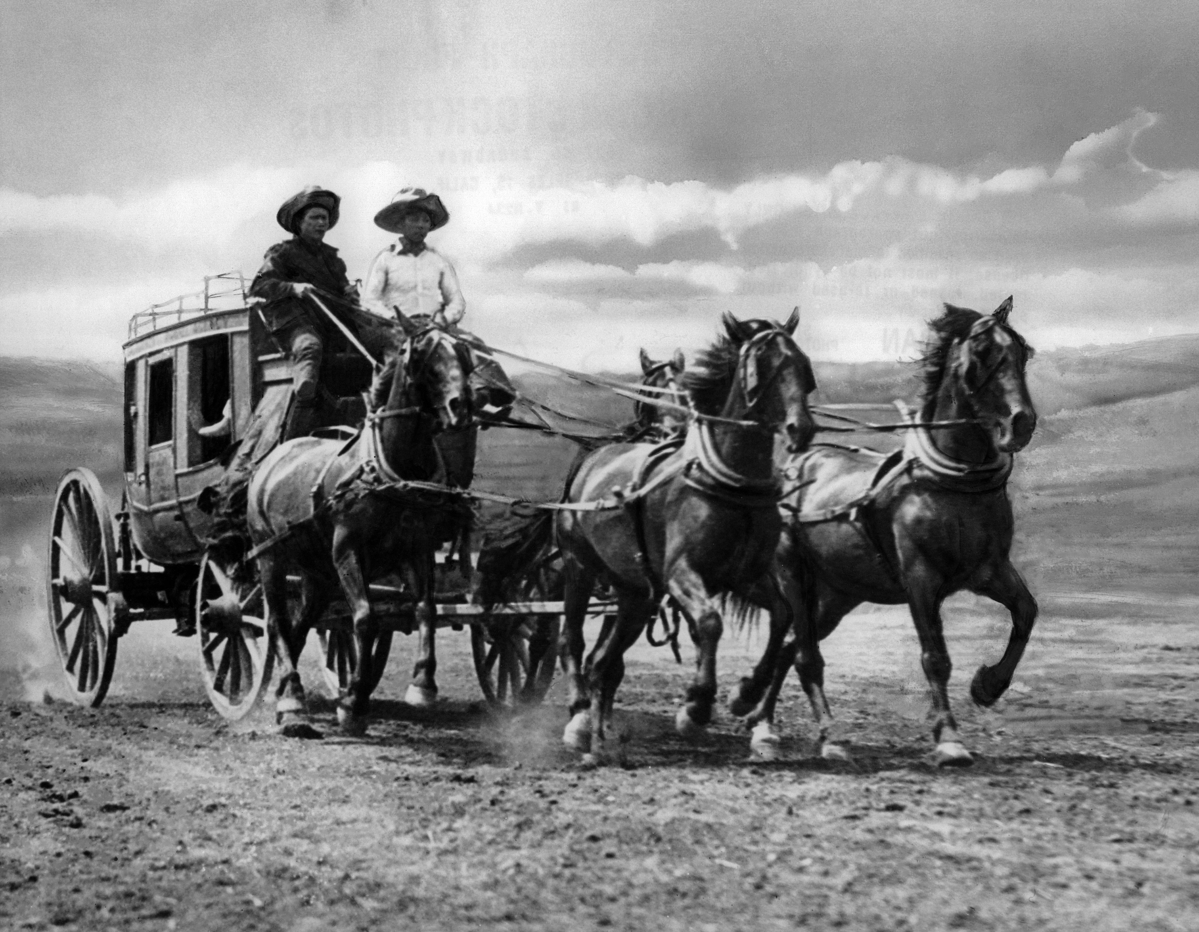 Black-and-white photo of a horse-drawn wagon with two drivers wearing wide-brim hats, pulled by several horses across an open dusty plain under a cloudy sky.