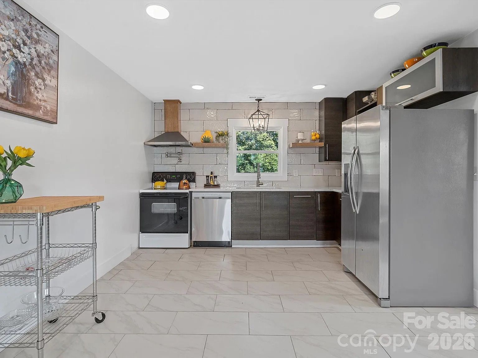 Modern kitchen with dark brown cabinets, stainless steel appliances, a window above the sink, white tiled floor and backsplash, and a metal rolling cart with yellow flowers.